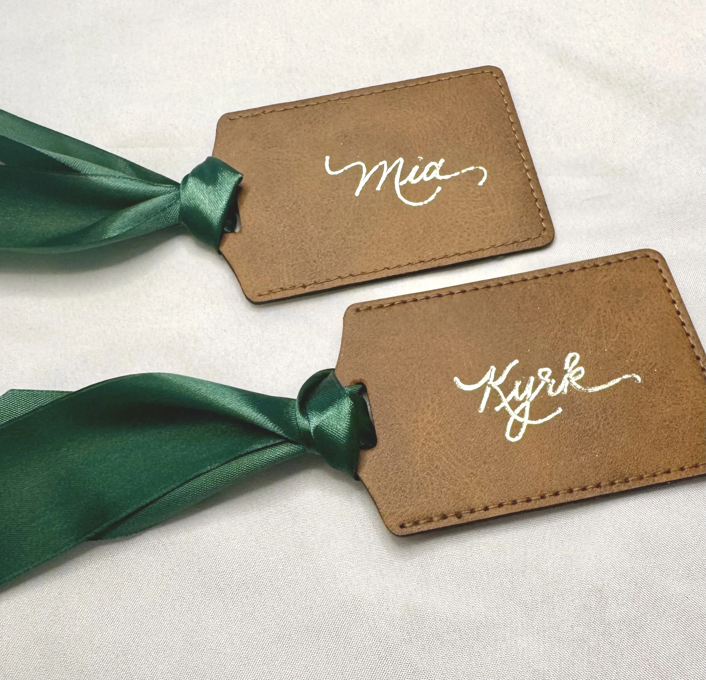 Two brown leather luggage tags with white cursive writing and attached green satin ribbons on a white fabric background.