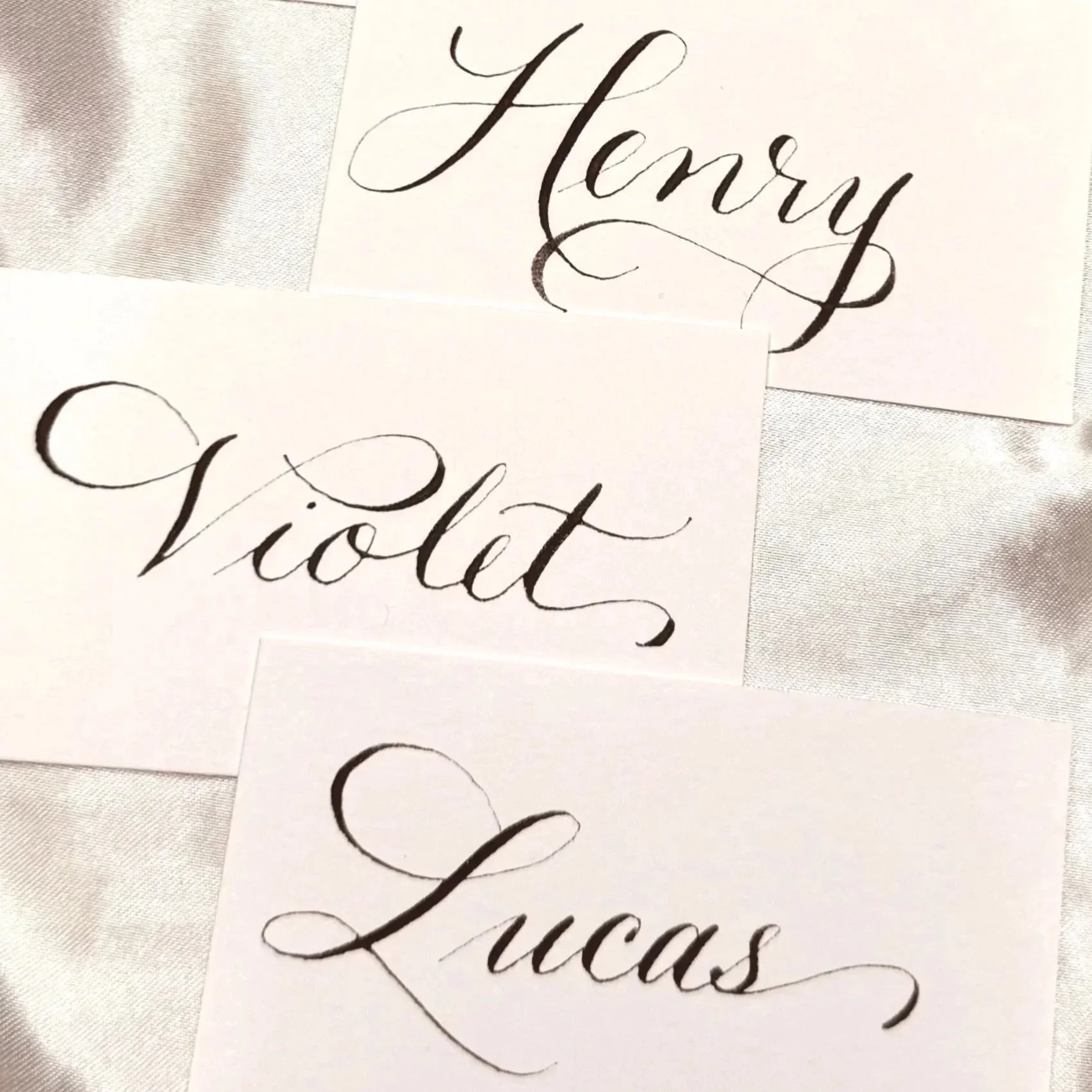 Three pieces of paper with elegant calligraphy, each displaying a different word: 'Harry,' 'Violet,' and 'Lucas,' on a silver fabric background.