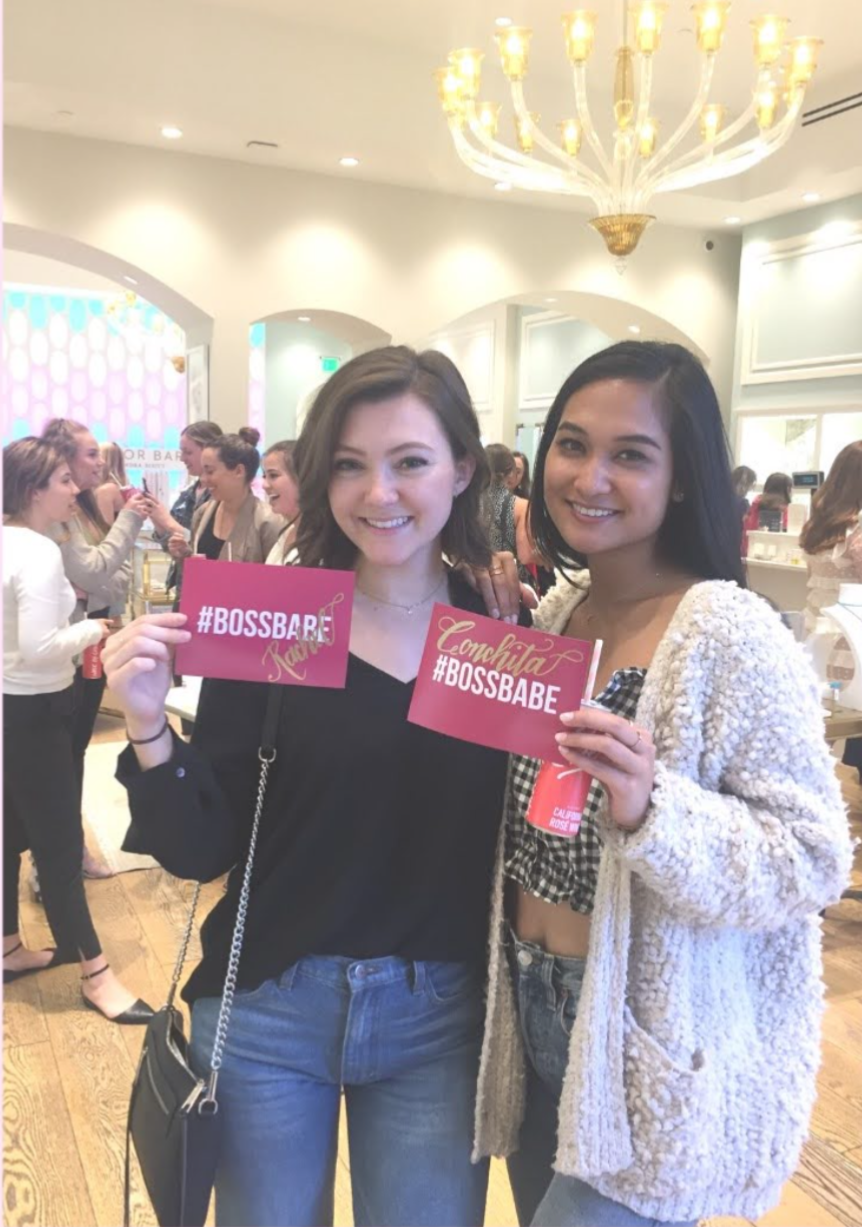 Two young women smiling and holding signs that read '#BOSSBABE' at an indoor event with many people in the background, softly lit with a chandelier overhead.