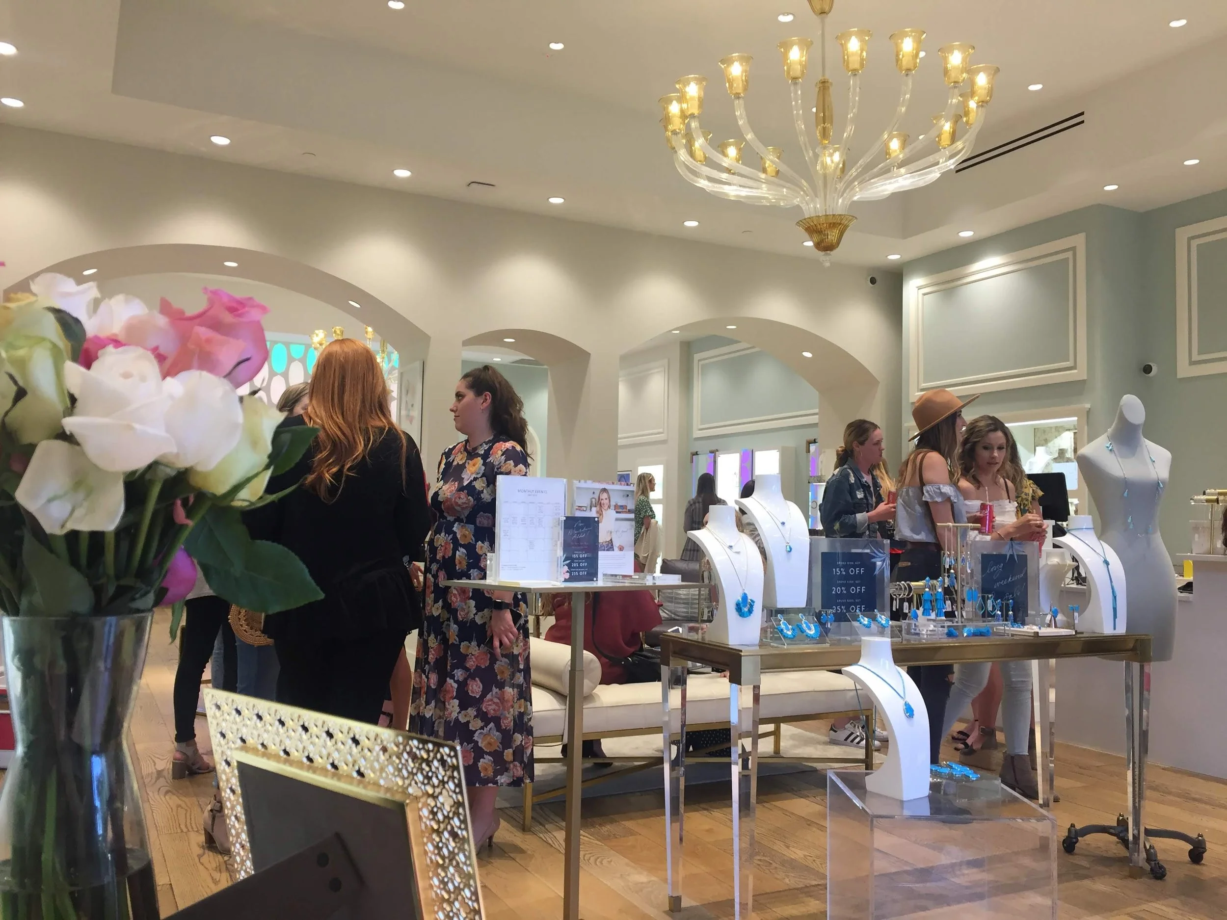 Inside a jewelry store with display cases and mannequins showcasing necklaces and earrings. Several women are shopping and talking, with a large chandelier and light blue walls in the background.