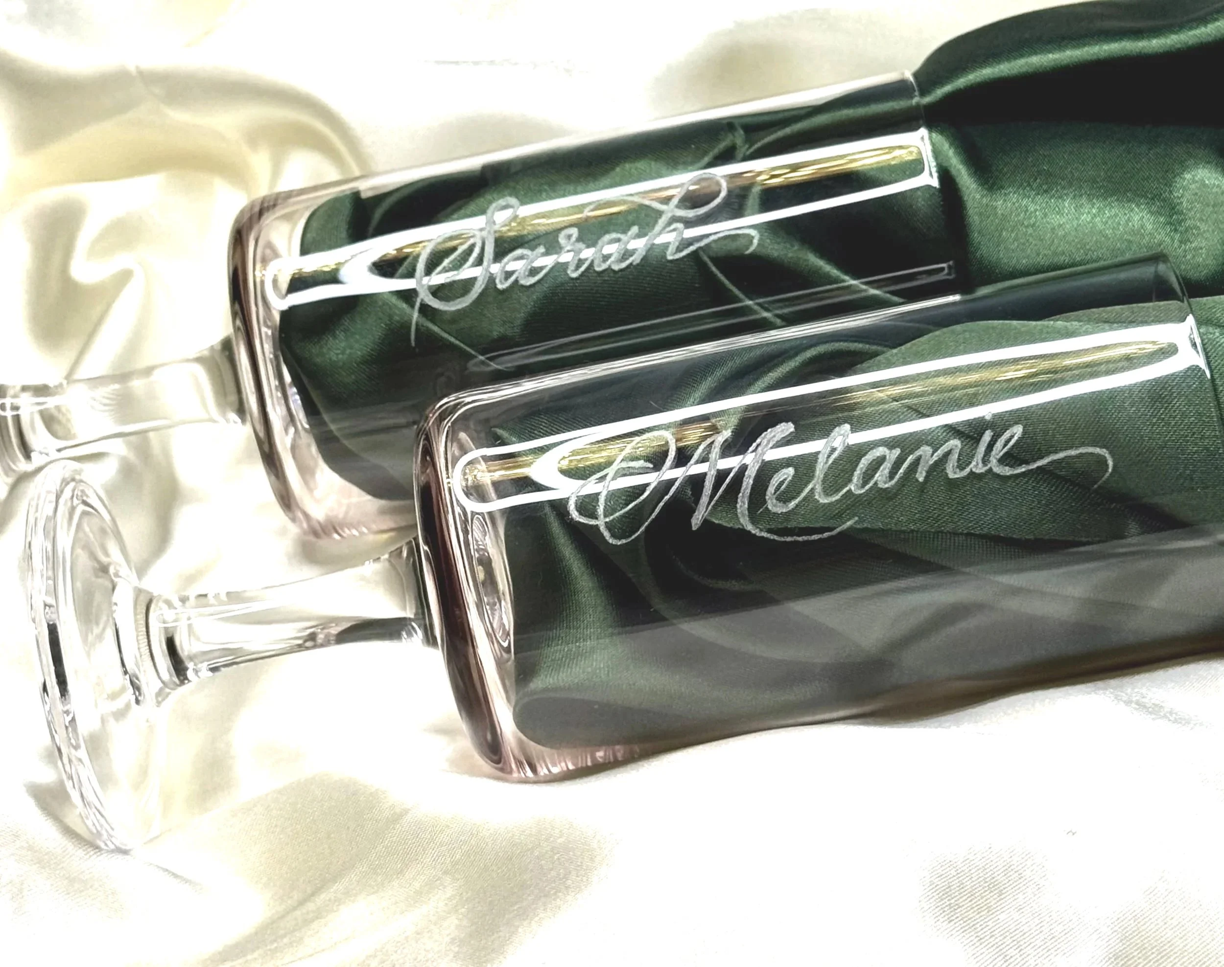 Two champagne flutes with engraved names 'Sarah' and 'Melanie' resting on a satin cloth, with a green gift box lined with green fabric.