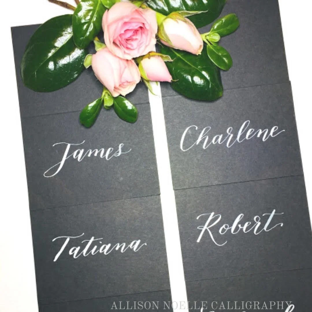 Place card with a pink rose and greenery on top, with four names written in white cursive font: James, Tatiana, Charlene, Robert.
