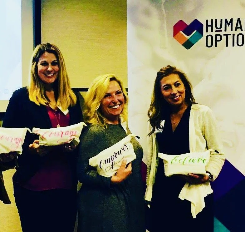 Three women smiling and holding white cloths with words written on them, standing in front of a backdrop that says 'HUMAN OPT.'