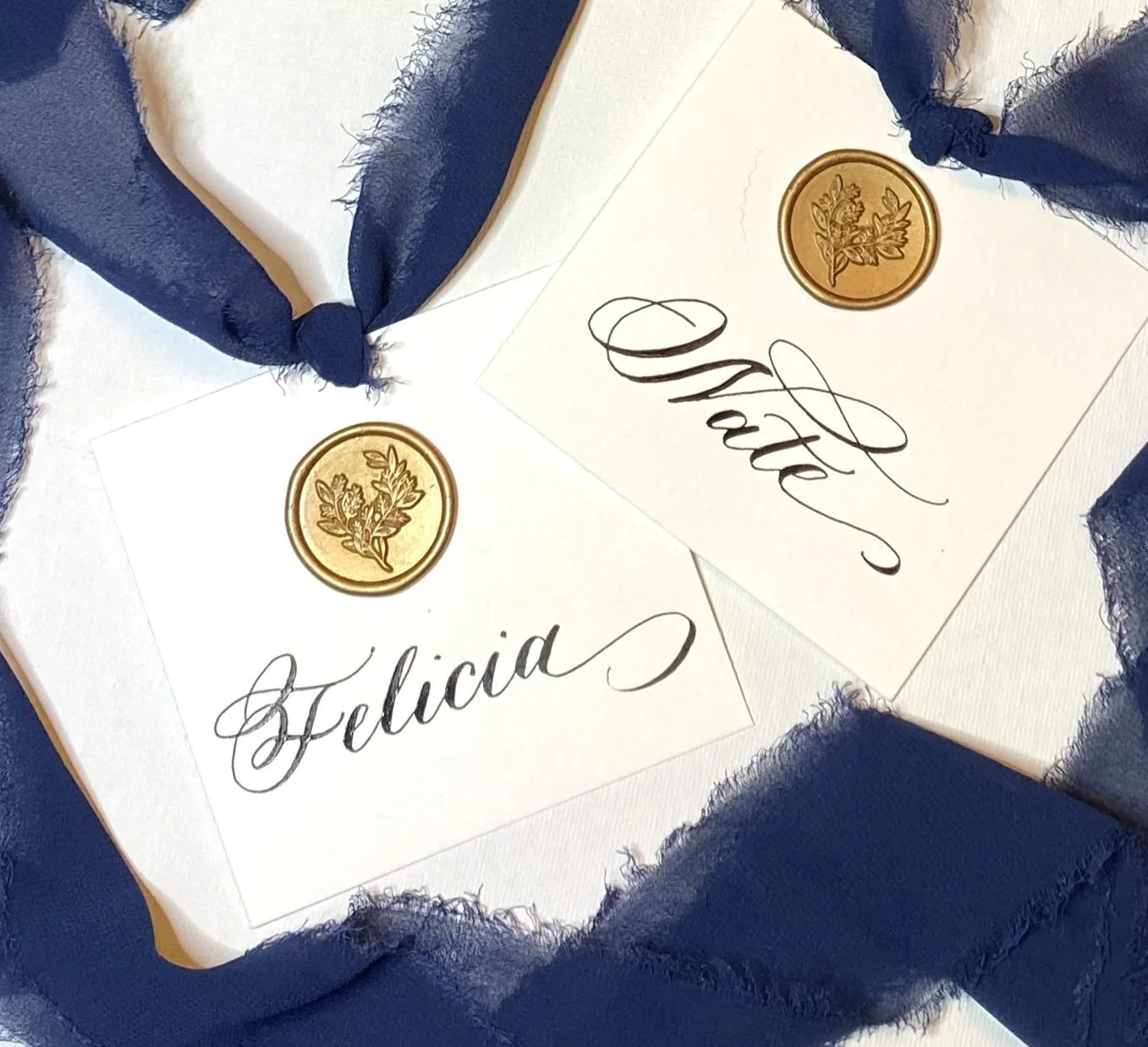 Two white cards with gold wax seals and the names 'Felicia' and 'Shai' written in elegant black calligraphy, surrounded by navy blue fabric with frayed edges.