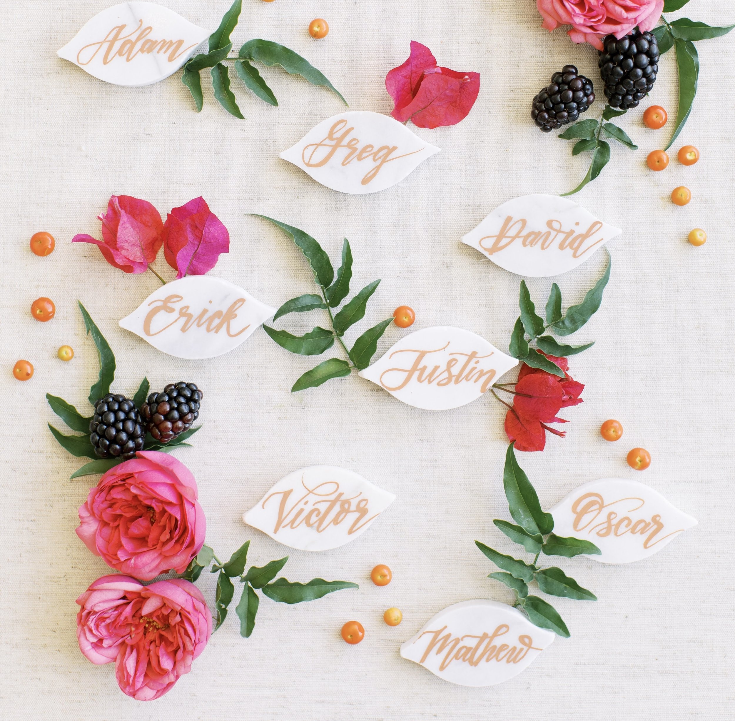 Decorative floral arrangement with pink roses, blackberries, small orange berries, green leaves, and white paper labels with peach-colored calligraphy names. The background is a light fabric.