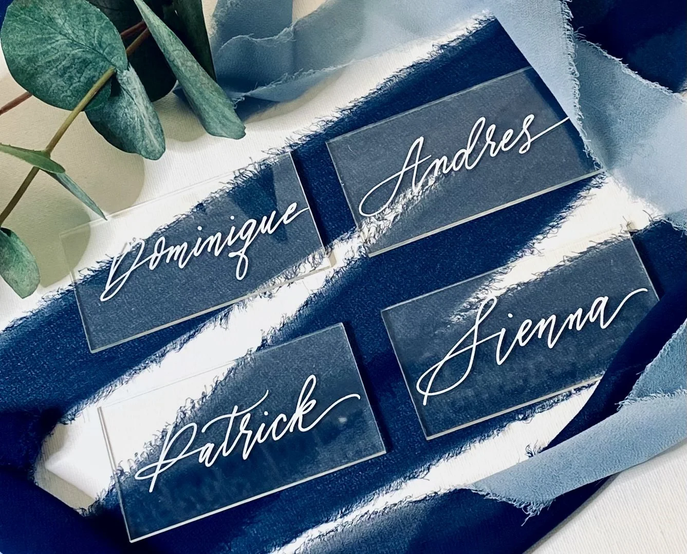 A wedding seating chart with clear acrylic name cards displaying names written in cursive: Dominique Andres, Patrick Sienna. The chart is decorated with blue frayed fabric and greenery.