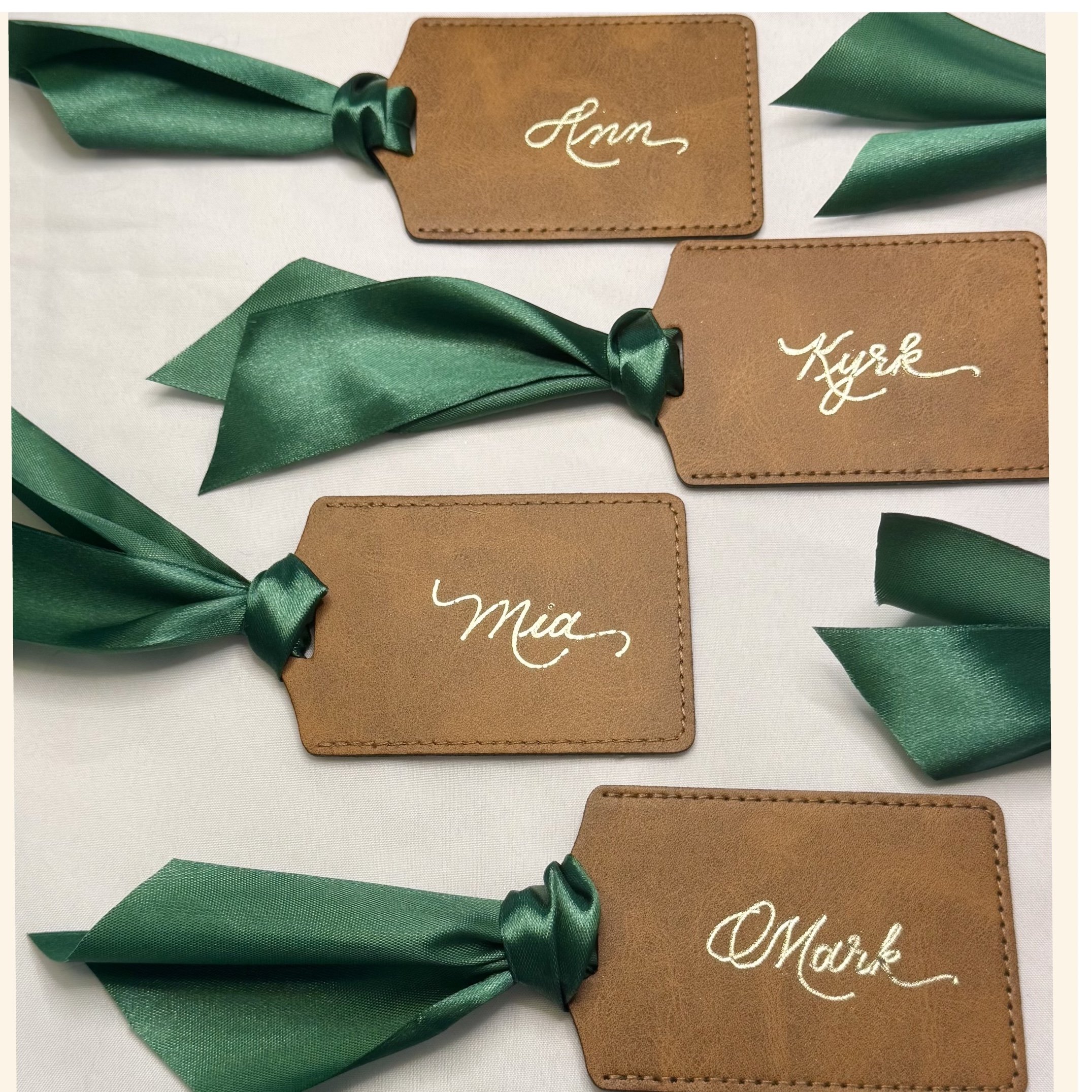 Close-up of brown leather tags with white cursive writing and green satin ribbons, arranged on a white surface.