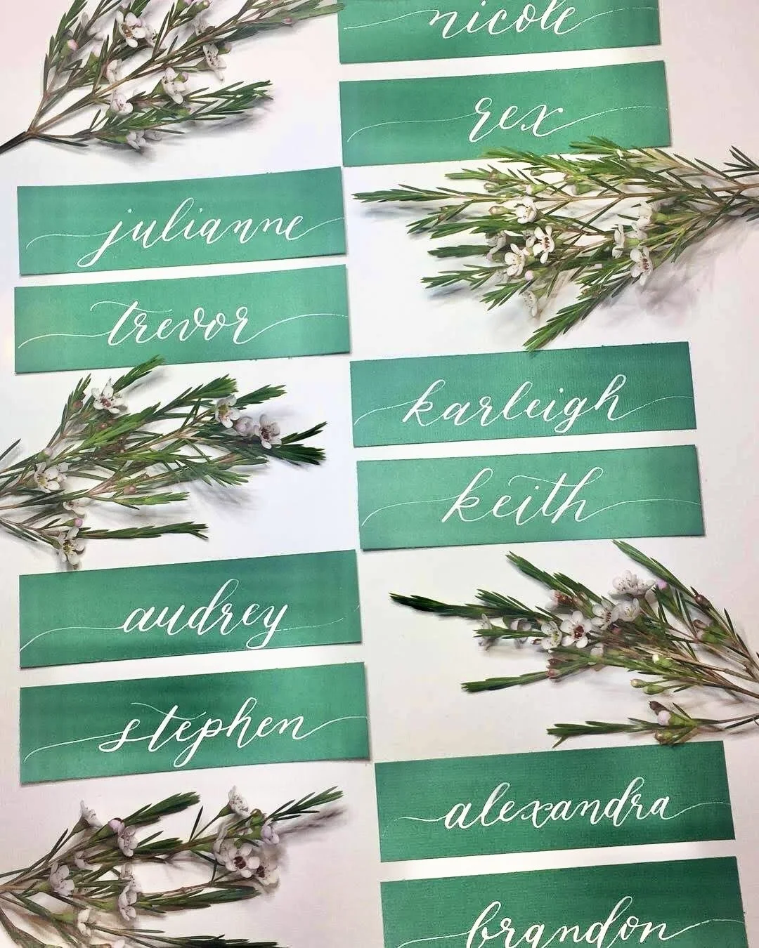 Name tags for a wedding or event, written in white cursive font on green backgrounds, arranged with sprigs of small white flowers and green leaves.