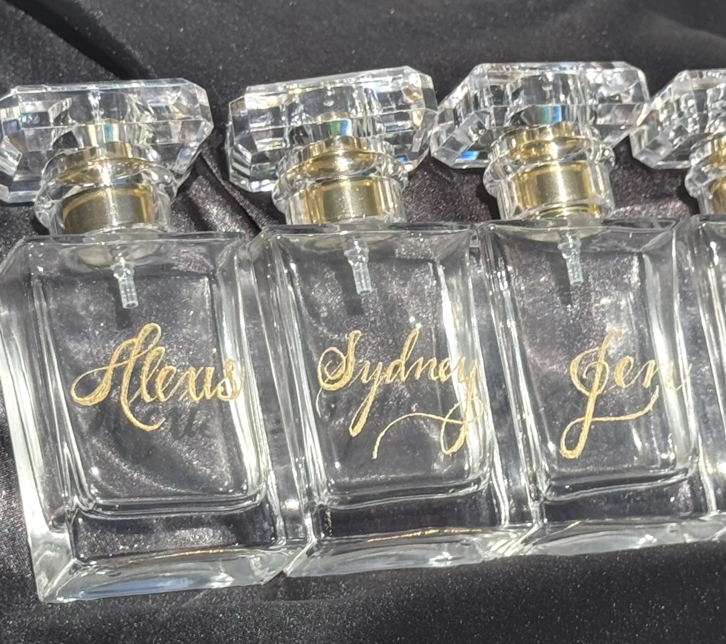 Three glass perfume bottles with gold writing and clear caps, labeled 'Alexis', 'Sydney', and '?', arranged on a black surface.