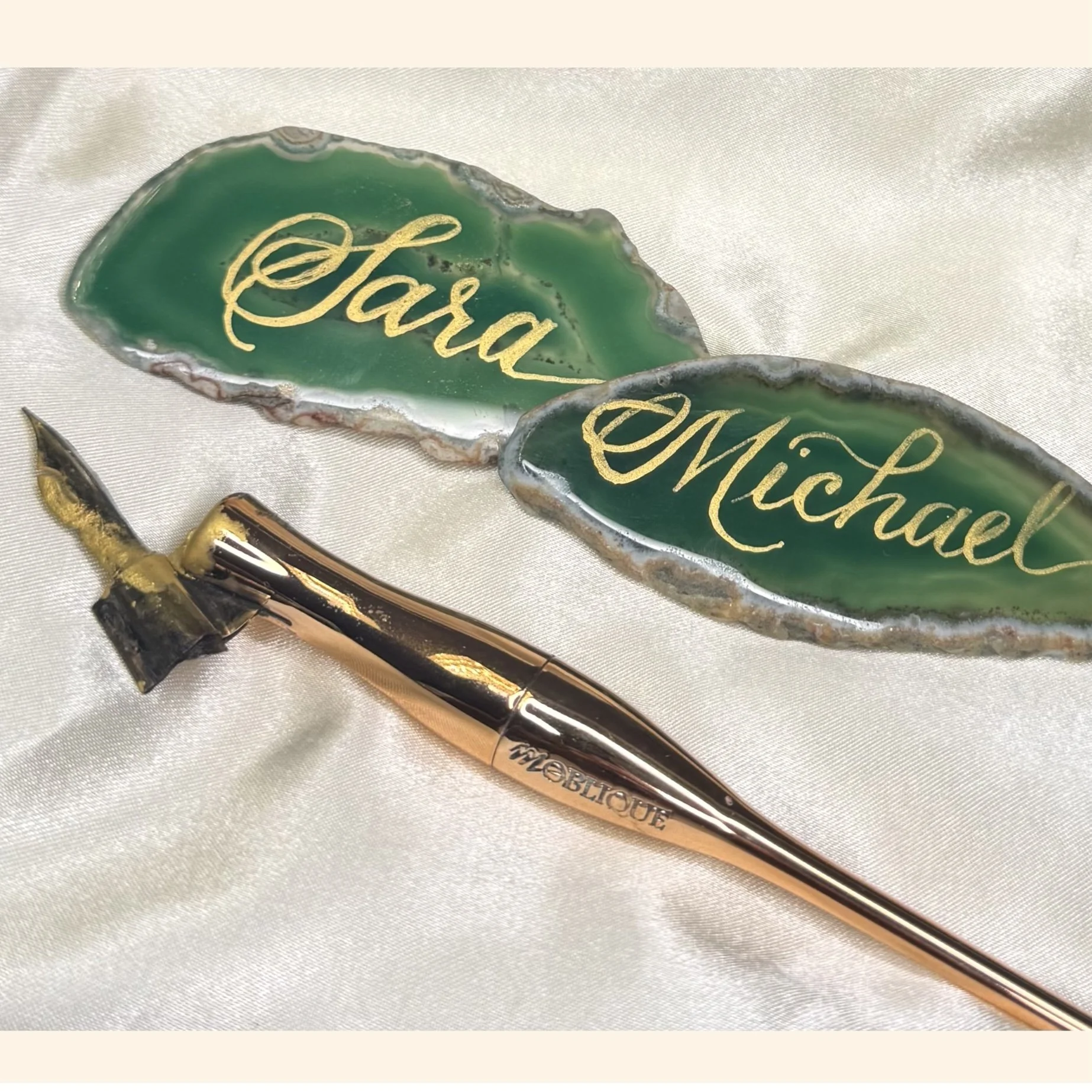 Decorative green agate stone with gold cursive text reading 'Lore' and 'Michael.' A gold and black pen is in the foreground, and the items are resting on a satin fabric.