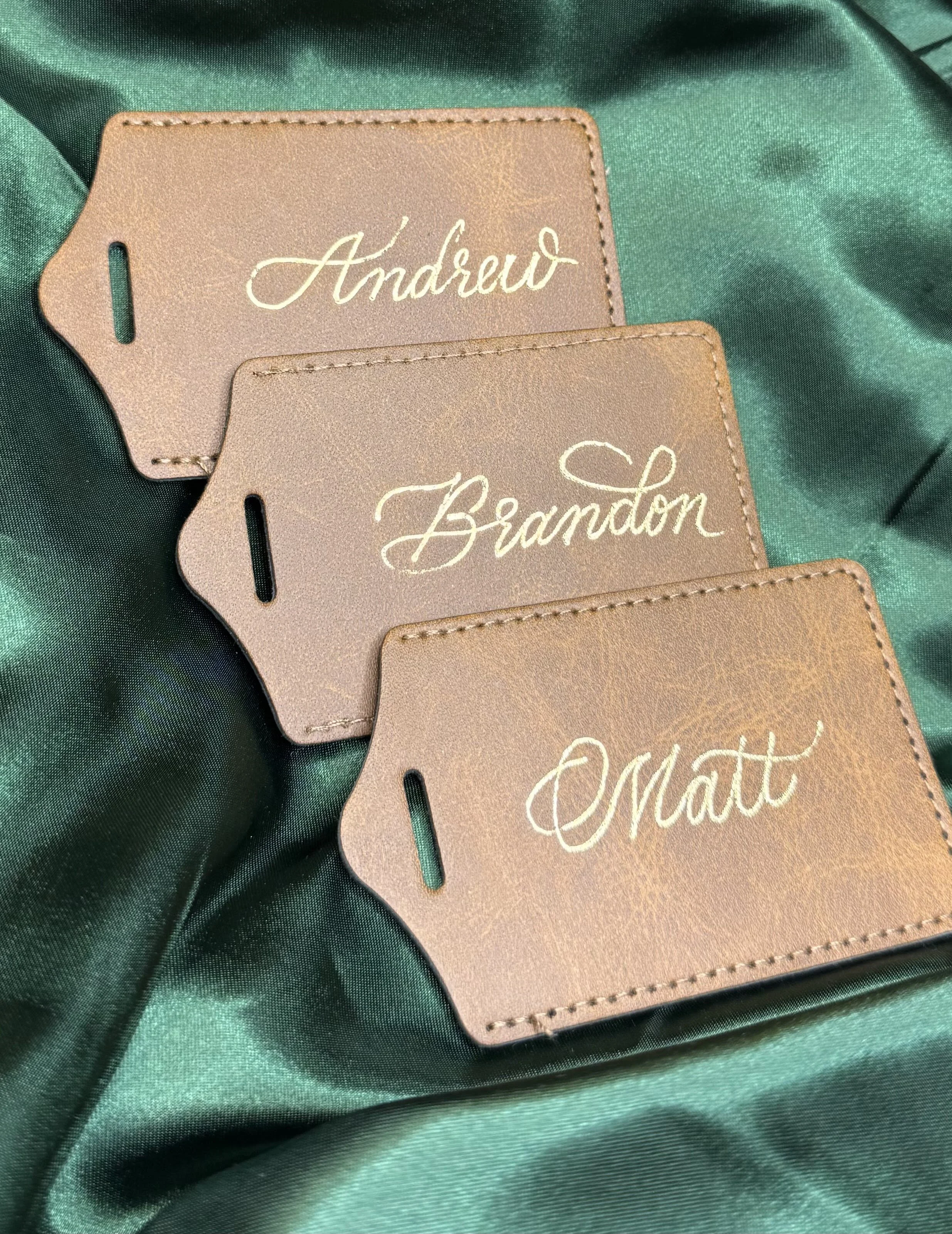 Three brown leather luggage tags with white cursive writing of the names Andrew, Brandon, and Matt, placed on dark green fabric.