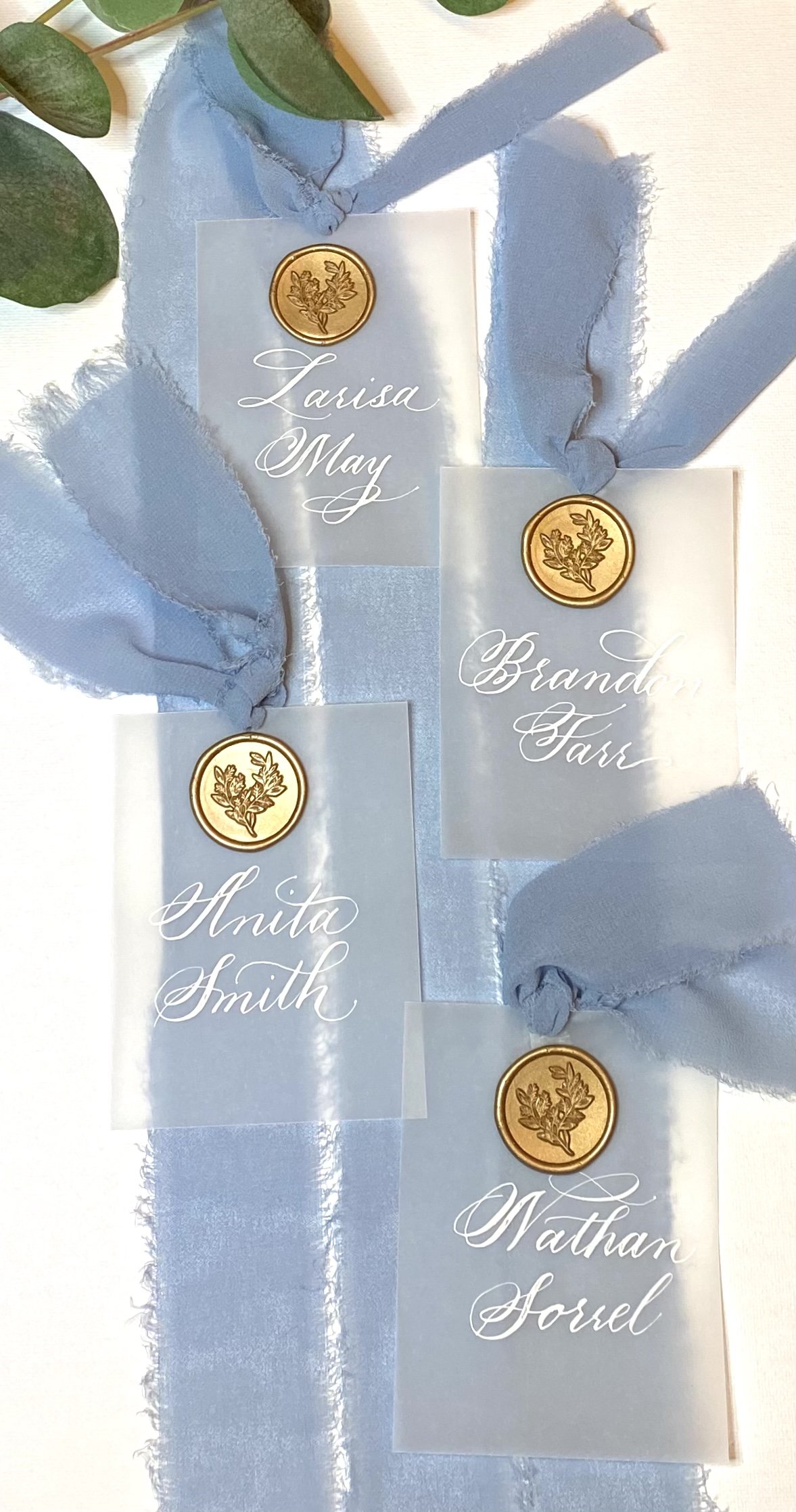Four wedding place cards with gold wax seals, names written in white cursive, on a light blue ribbon with greenery leaves nearby.