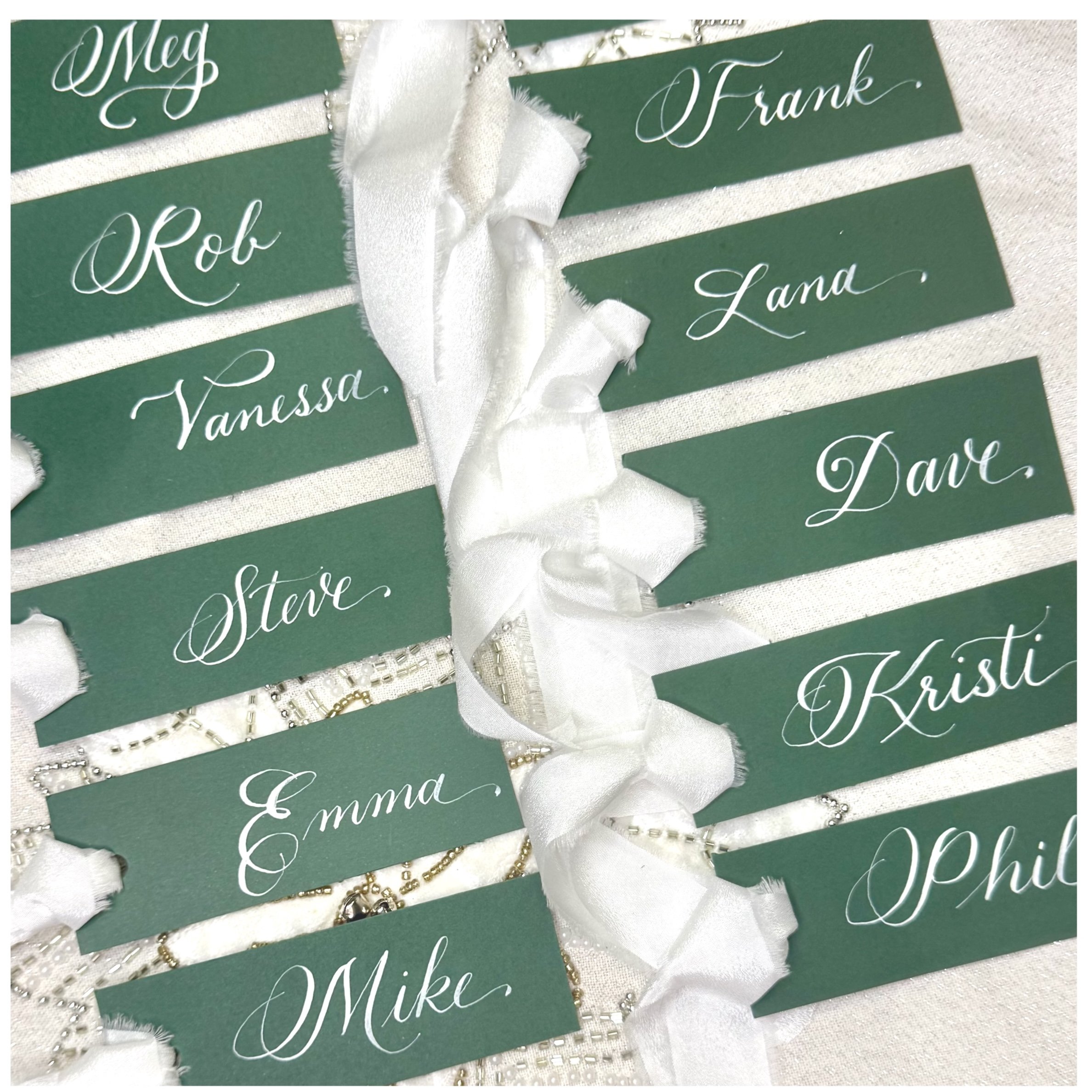 A collection of green name cards with white cursive writing, each featuring a different name such as Meg, Frank, Rob, Vanessa, Steve, Emma, Mike, and Kris, decorated with white satin ribbons, pearls, and beaded embellishments on a white textured back
