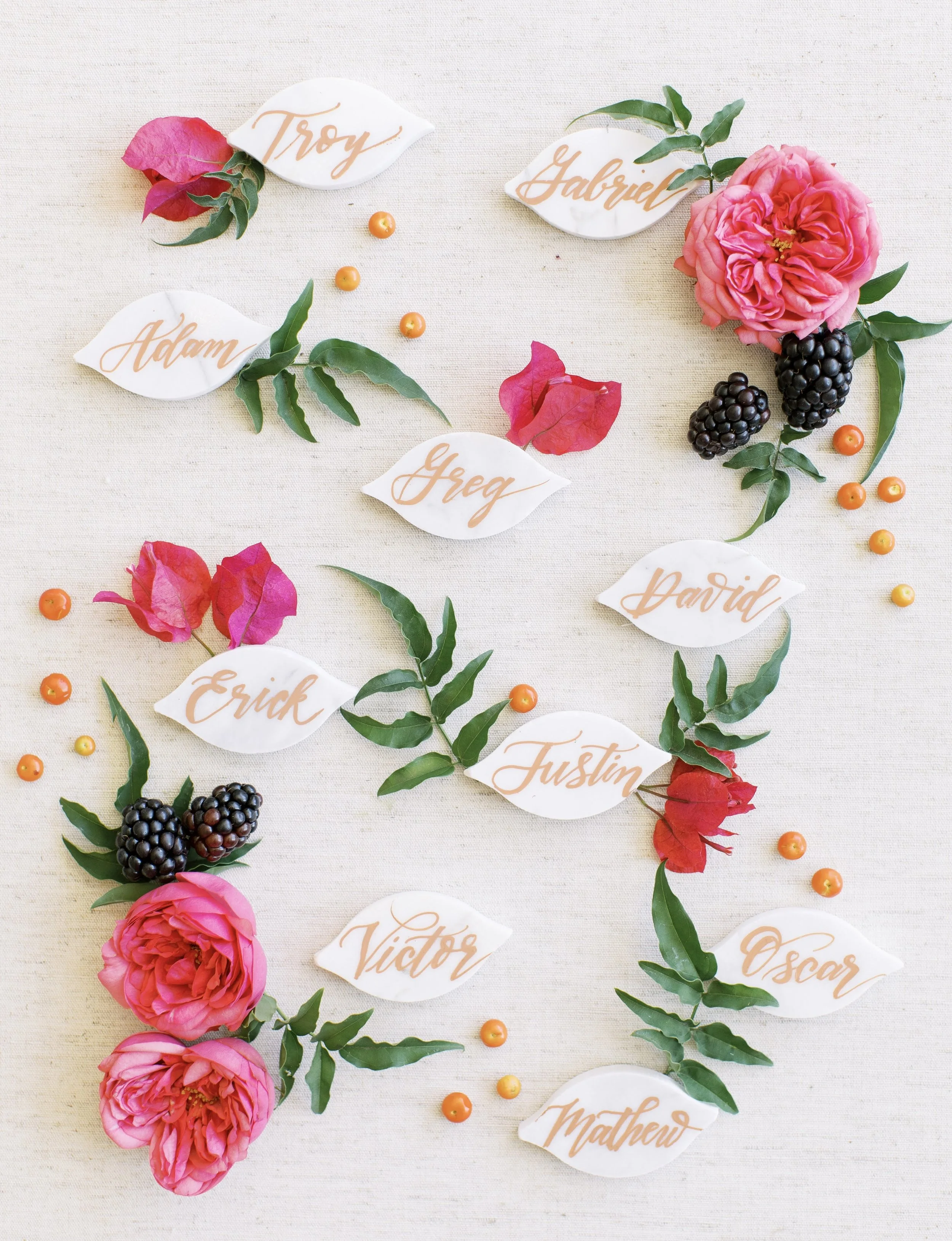 Decorative flat lay with pink roses, blackberries, orange berries, green leaves, and white paper leaves with orange calligraphy names on a light beige fabric background.