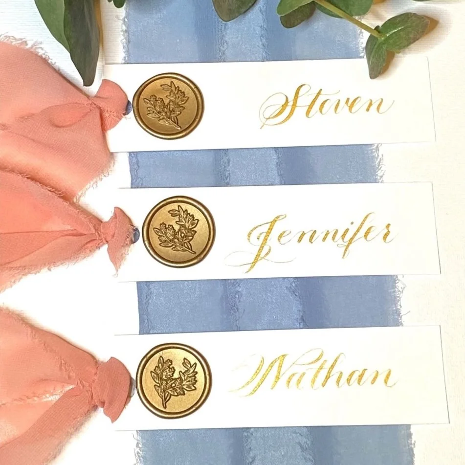 Three place cards with gold wax seals, gold calligraphy of names 'Stavn,' 'Jennifer,' and 'Nathan,' on a blue and white table runner, and pink fabric decorations.