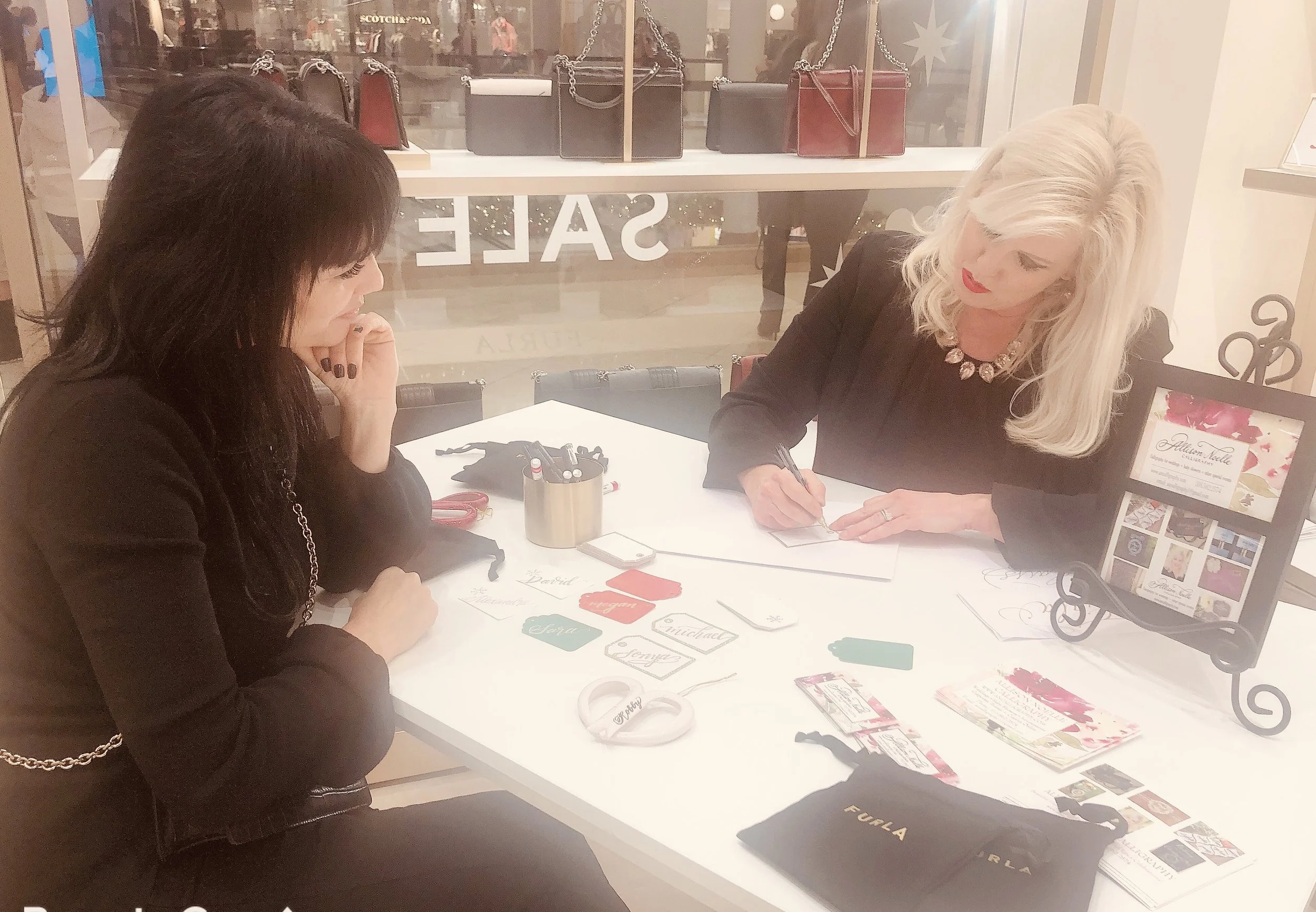 Two women sitting at a white table in a store, one with dark hair and the other with blonde hair, engaged in customizing or designing jewelry or cards, with various crafting supplies and promotional material on the table.