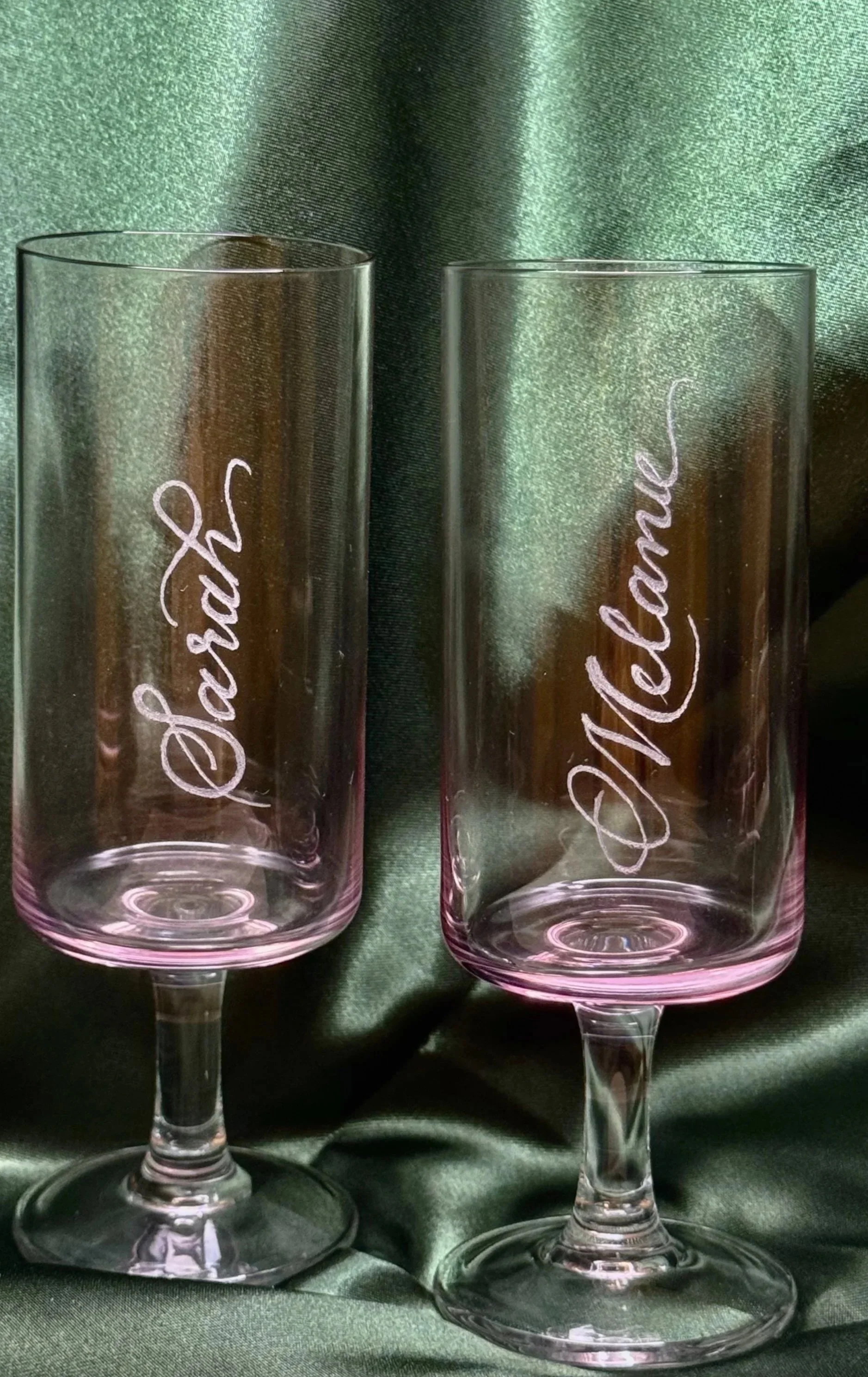Two transparent champagne flutes with pink tint at the base and pink inscriptions that read 'Dad' on the left and 'Mom' on the right, set against a dark green satin fabric background.