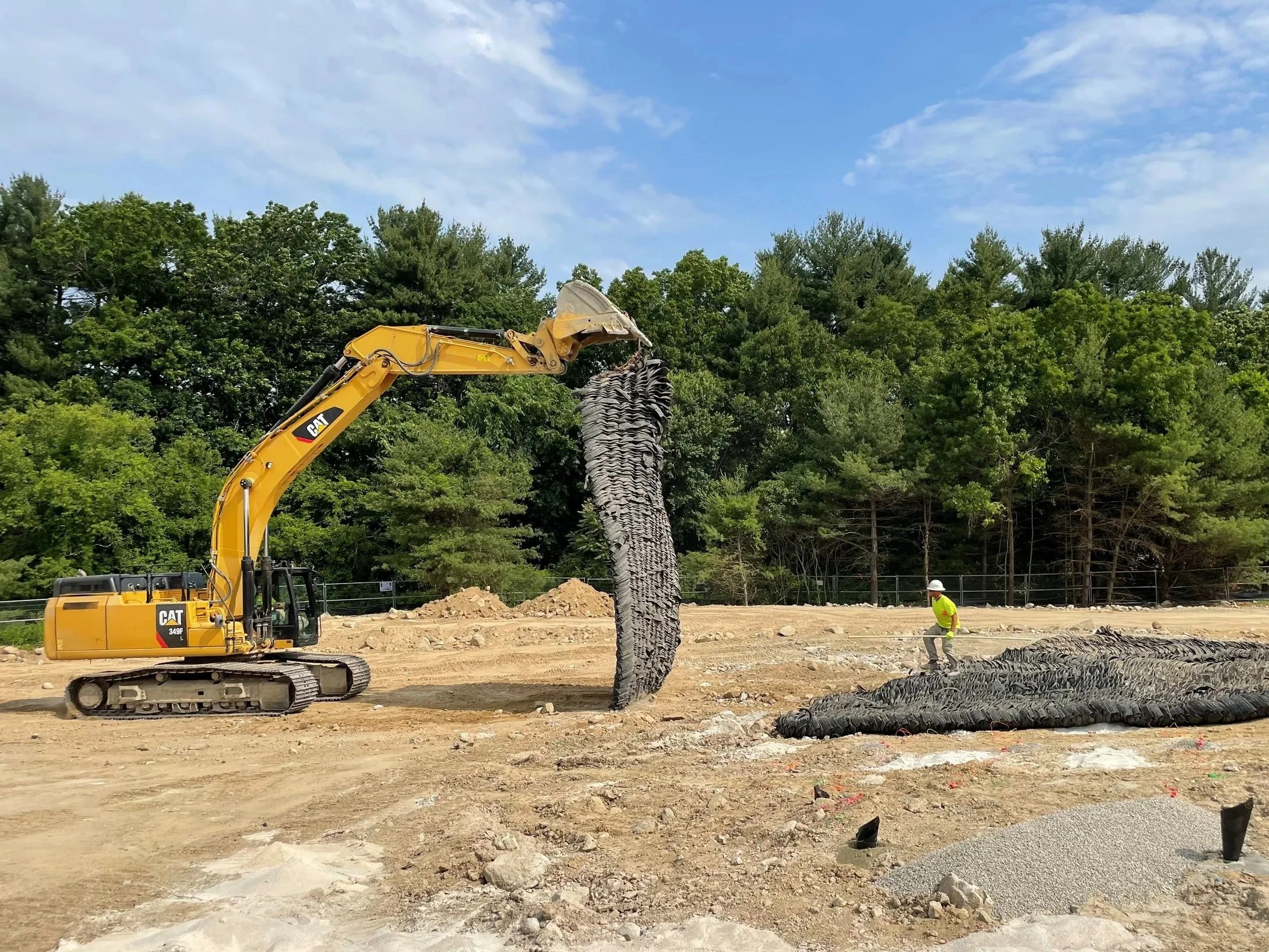 Construction site with a yellow CAT excavator lifting a large bundle of black tubing, worker in a yellow vest and white helmet standing nearby on dirt ground, surrounded by trees and blue sky.