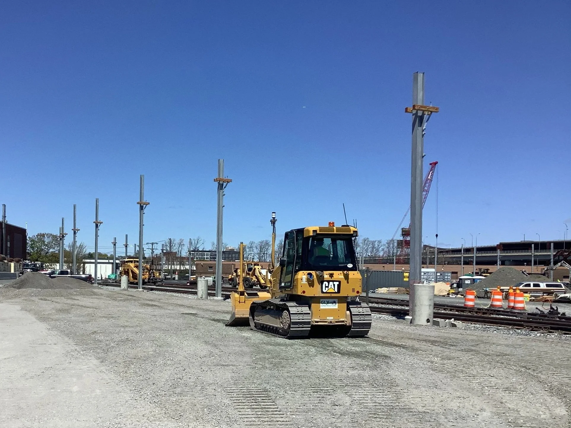 Construction site with a small CAT bulldozer on dirt ground, incomplete utility poles, and construction equipment in the background under a clear blue sky.