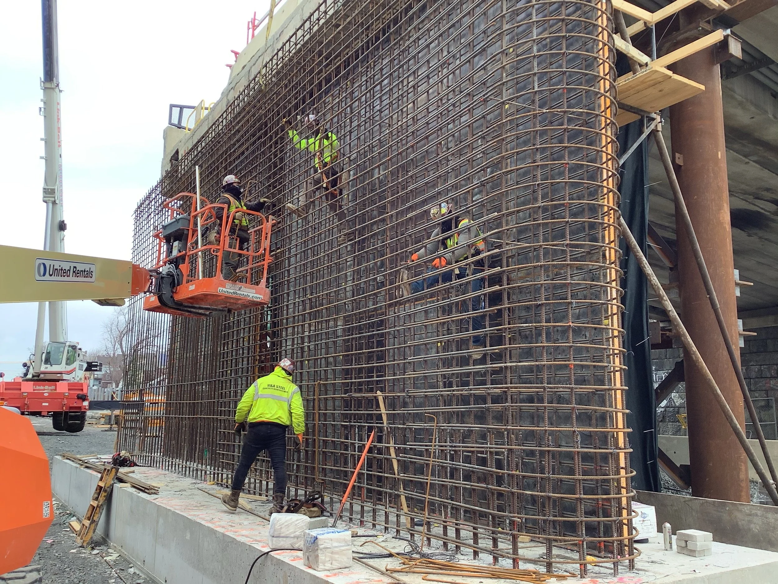 Construction workers installing rebar reinforcement on a concrete structure at a construction site, using a lift to reach higher areas.