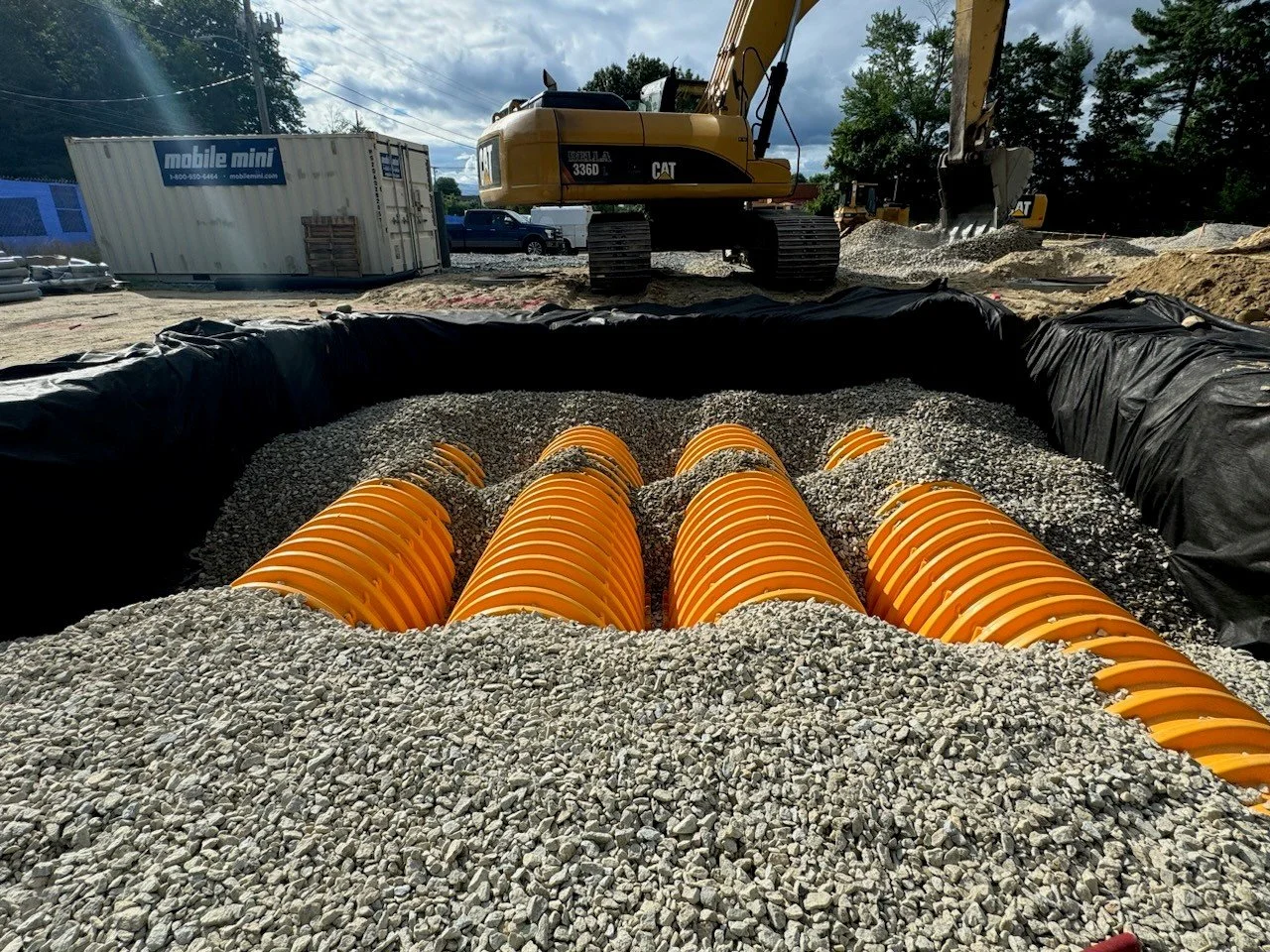 Construction site with a large excavator and orange drainage pipes installed in gravel, surrounded by black plastic sheeting, under a partly cloudy sky.
