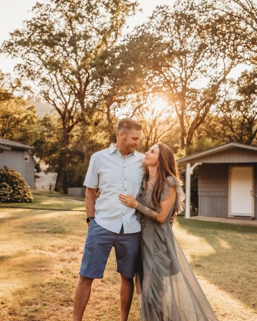 A couple standing outdoors at sunset, looking at each other, with trees and houses in the background.