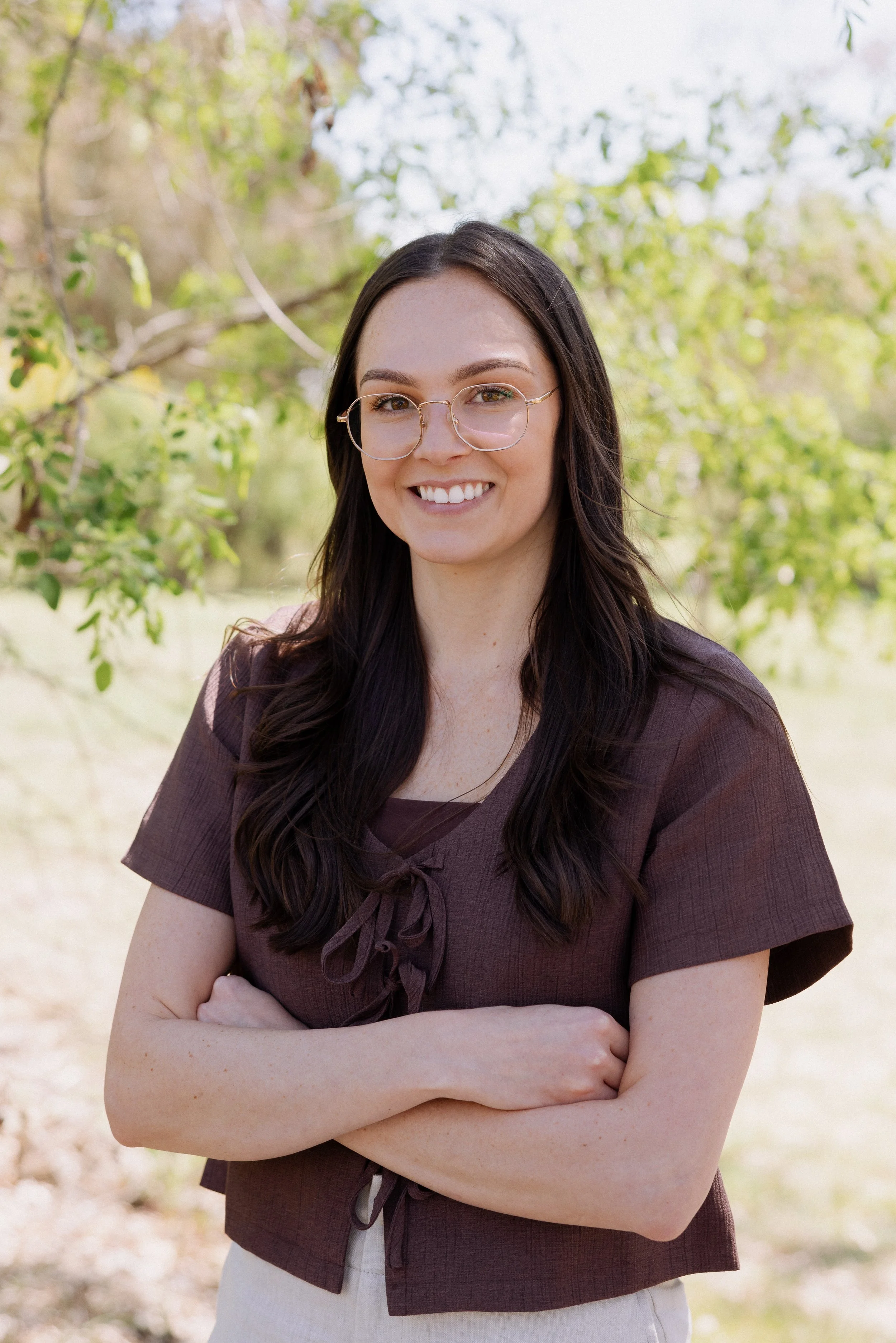 A young woman with long dark hair and glasses smiling outdoors, crossing her arms, in front of green trees.