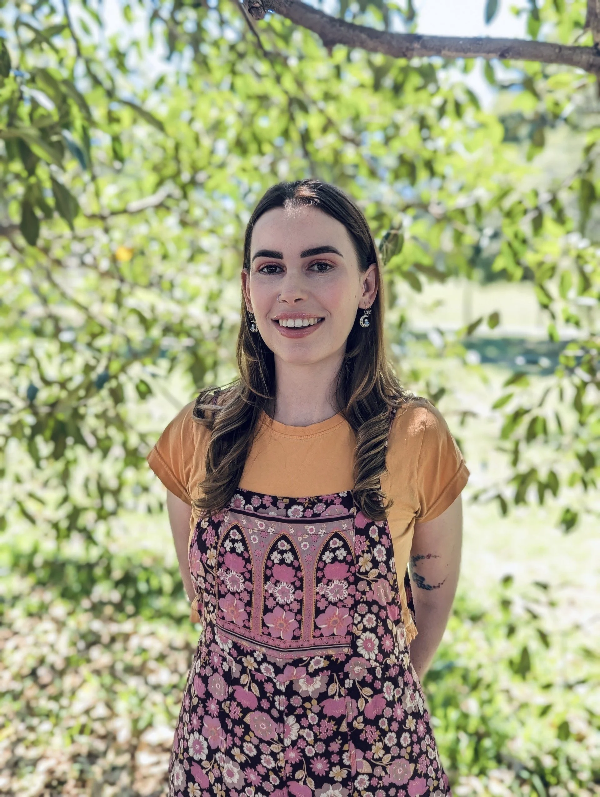 A young woman with long brown hair, smiling, standing outdoors with lush green trees in the background, wearing a patterned apron over an orange t-shirt.