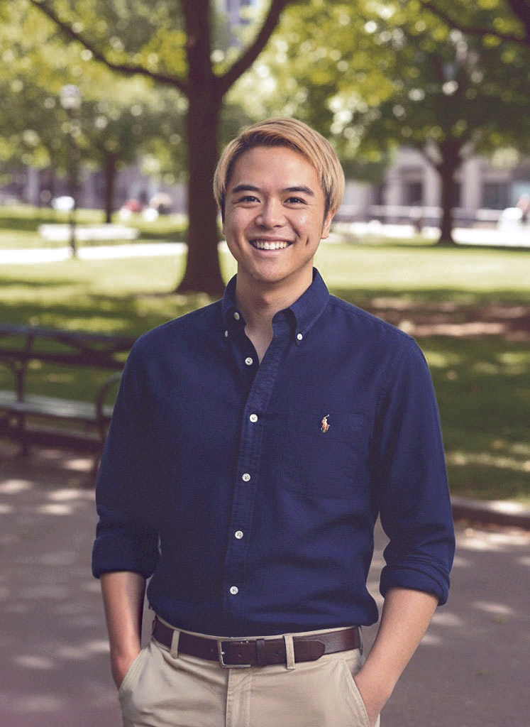 A young man with blond hair and a friendly smile, wearing a dark blue button-up shirt with rolled-up sleeves and khaki pants, standing outdoors in a park with trees and benches in the background.