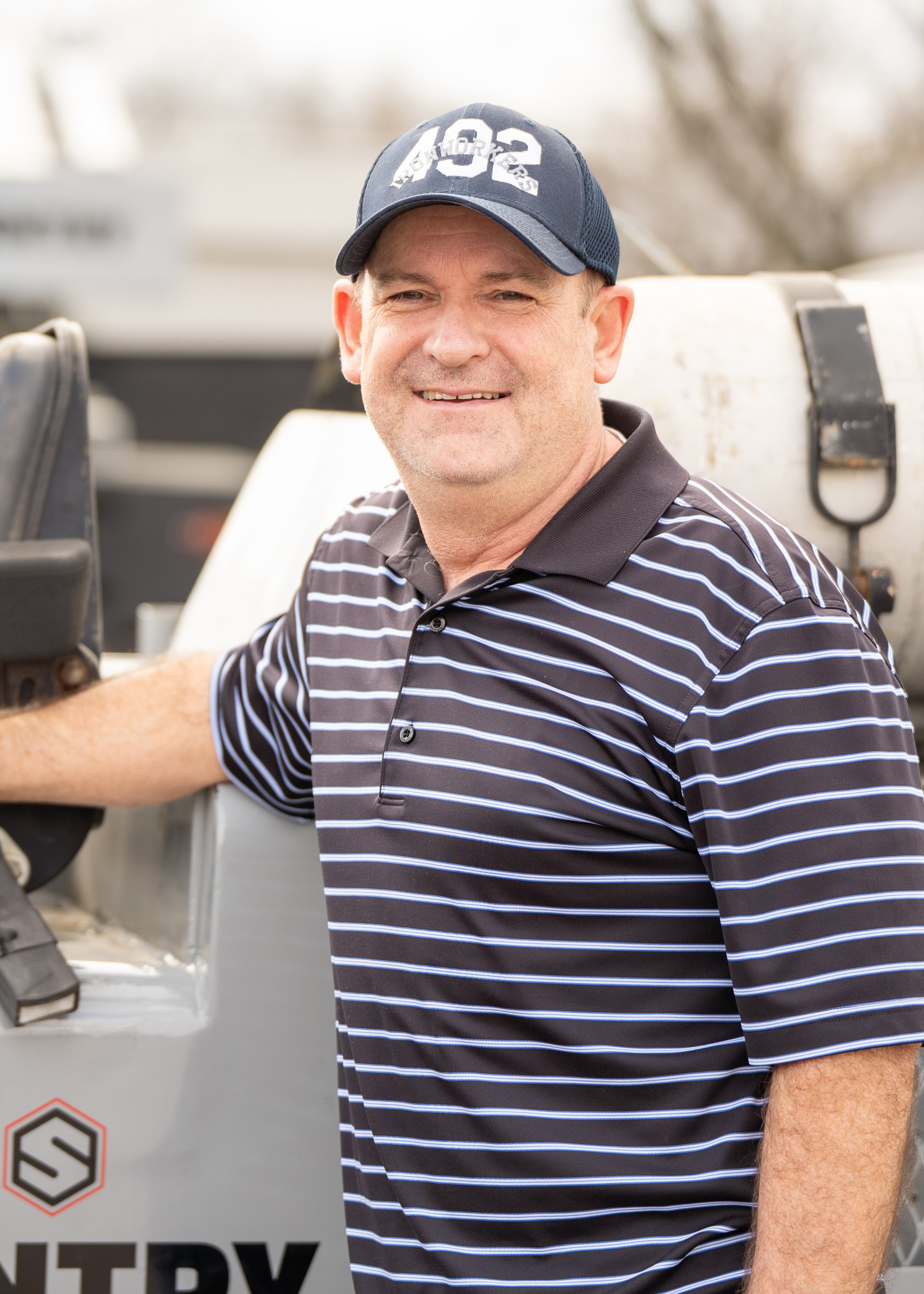A smiling man wearing a dark blue baseball cap and a black and white striped polo shirt, standing outdoors near machinery.