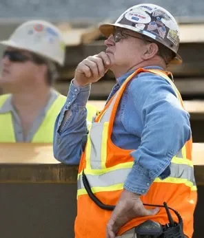Construction worker wearing a helmet, safety vest, and work clothes, with hand on chin, on a construction site.