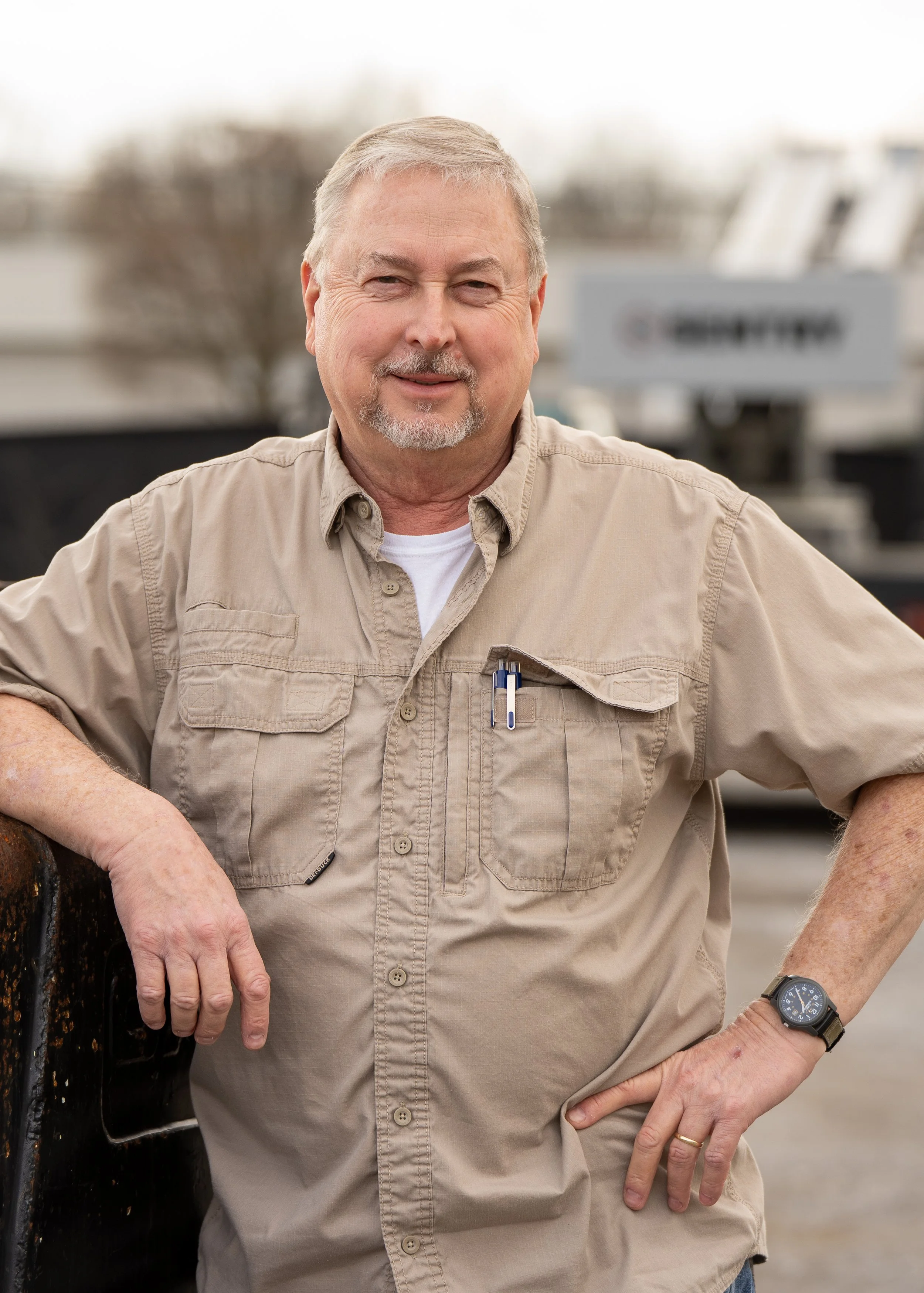 A middle-aged man with gray hair and a beard stands outdoors, smiling at the camera, wearing a beige button-up shirt, a black watch, and a ring, with blurred background of equipment and a sign.