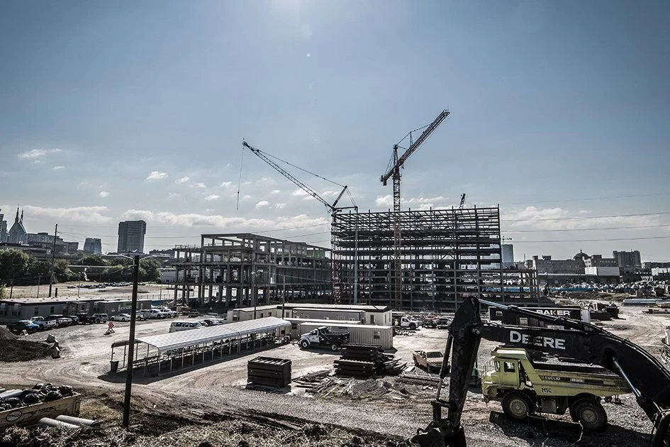 Construction site with a partially built multi-story building, cranes, construction vehicles, and equipment under a clear sky.