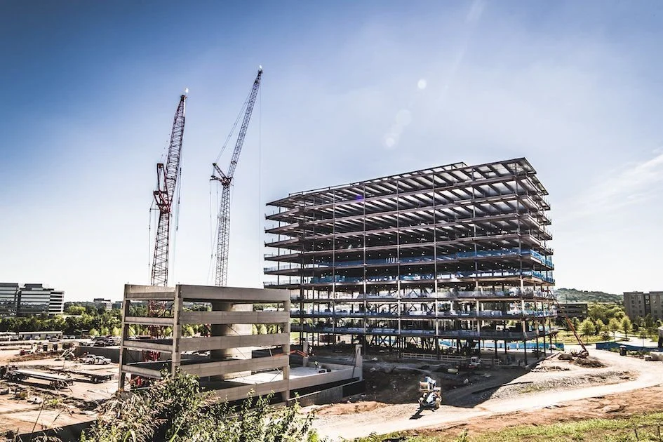 A multi-story building under construction with a steel frame structure, cranes nearby, and construction equipment on site.