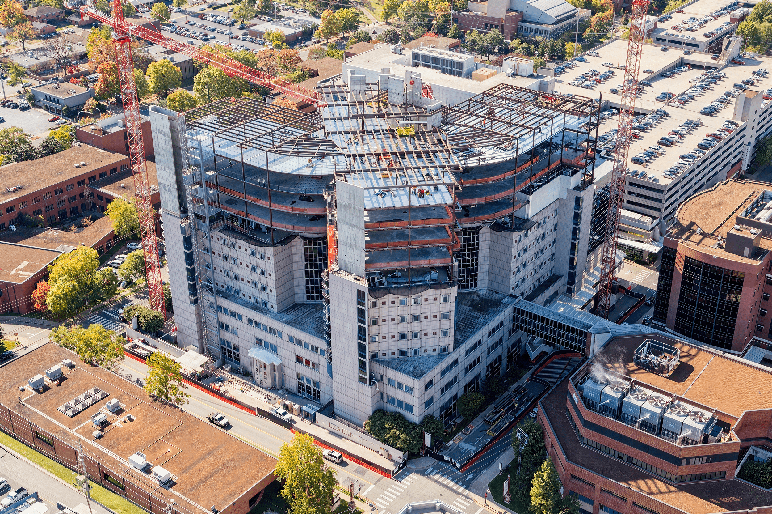 Aerial view of a tall commercial building under construction with two red cranes, surrounded by parking lots, roads, and other buildings with green trees.