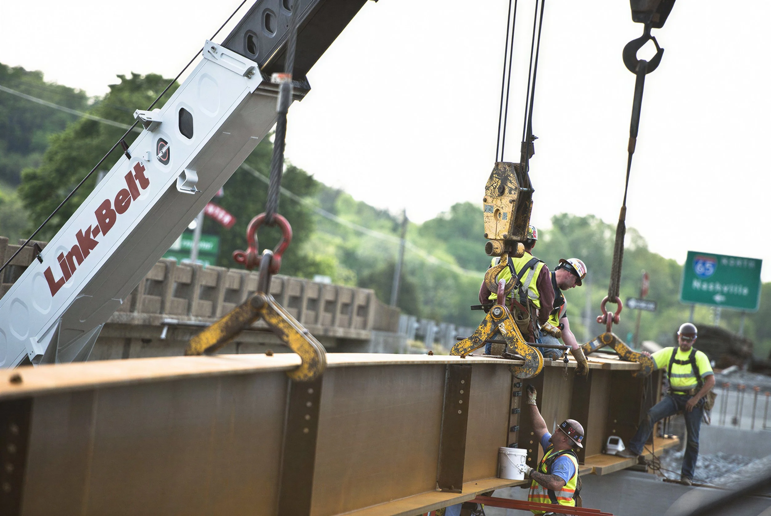 Construction workers wearing safety gear work on a bridge using a crane labeled 'Linde-Belt' to position large metal beams.