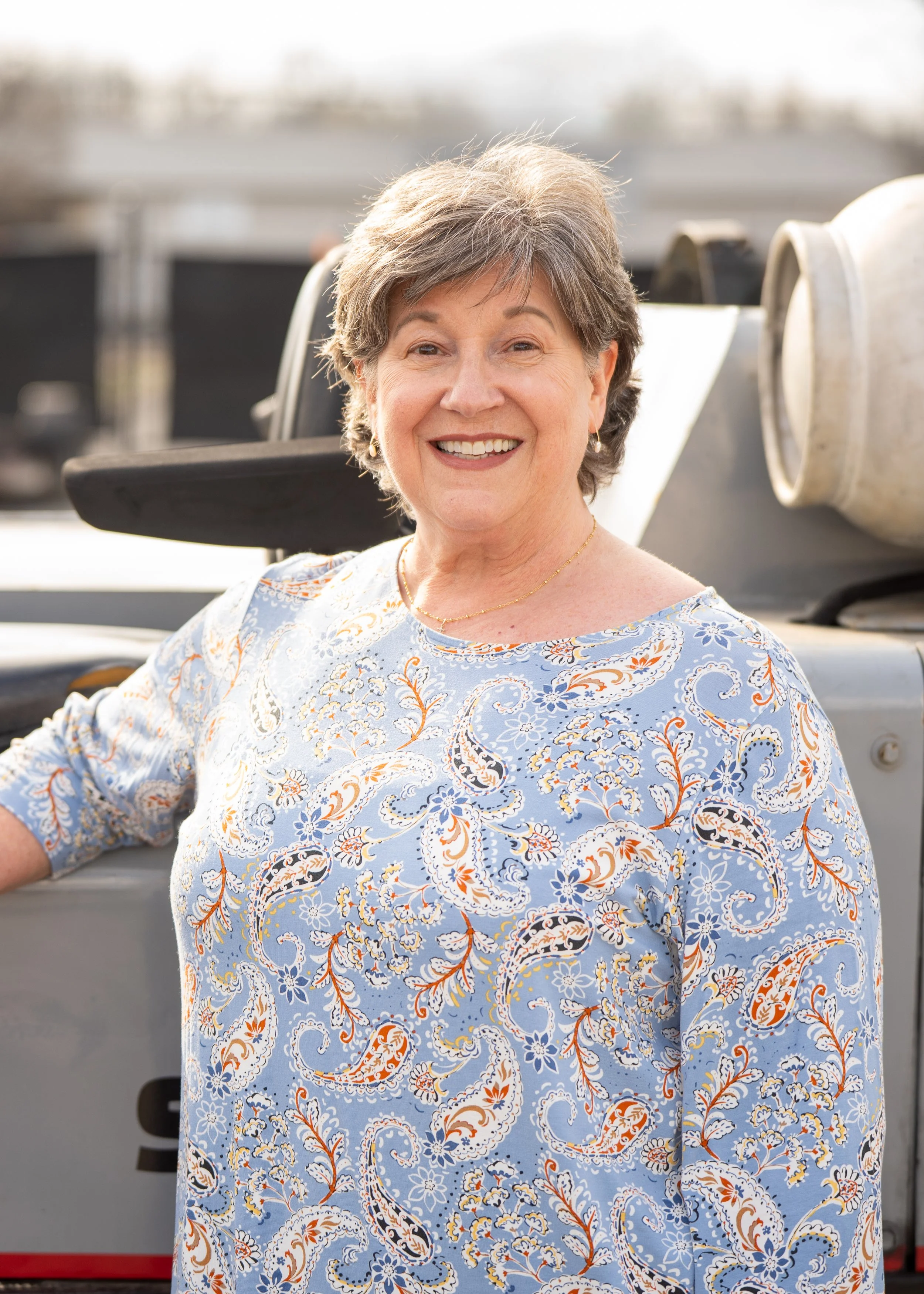 A smiling elderly woman with short gray hair, wearing a light blue dress with an orange and white floral pattern, standing in front of a gray vehicle with blurred background.