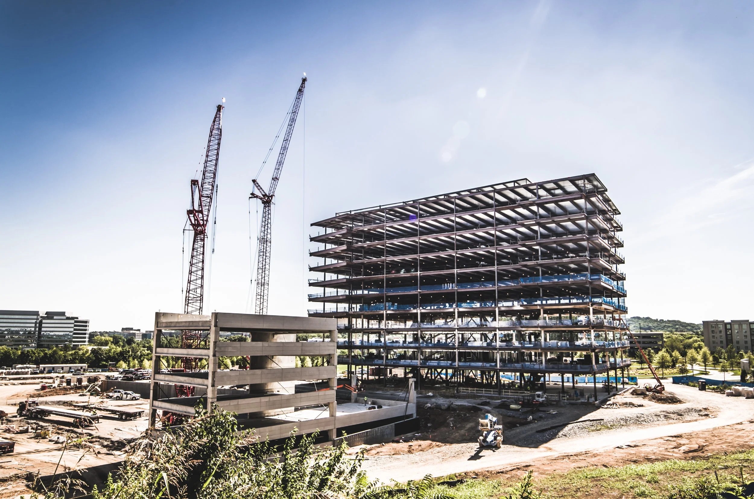 Construction site with a multi-story building framework, two cranes, and a partly built parking structure against a clear blue sky.