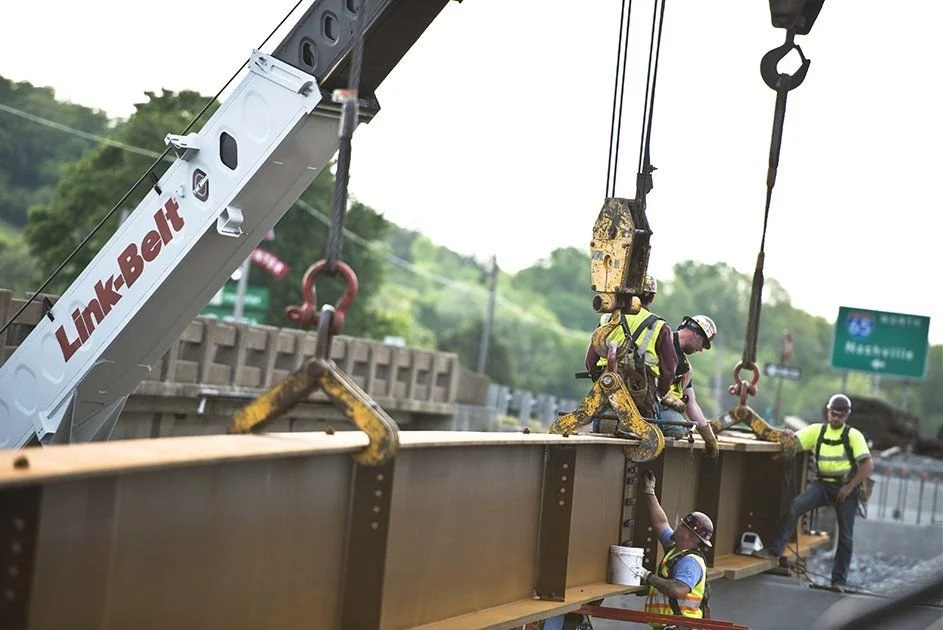 Construction workers on a highway with a crane lifting a steel beam