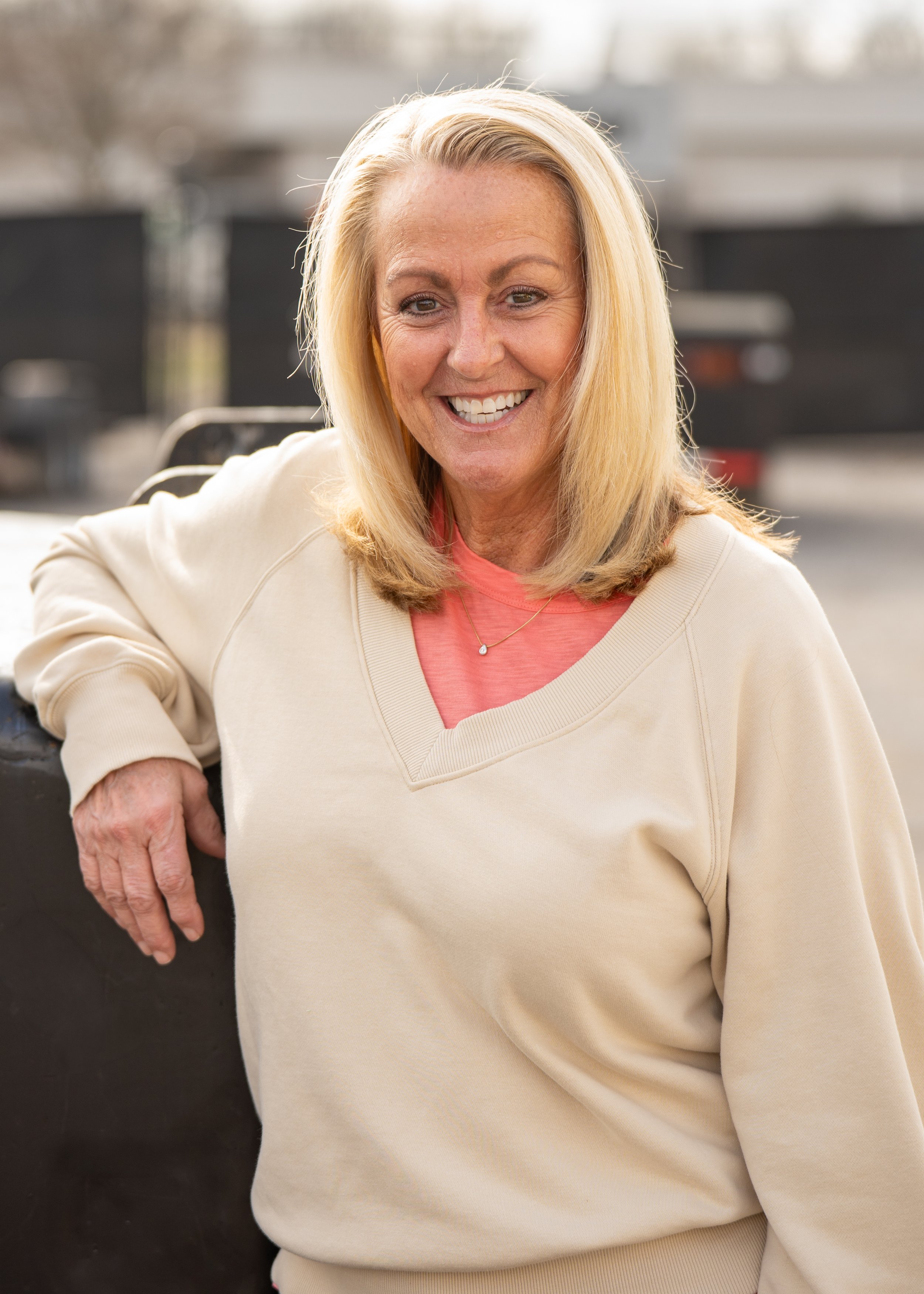 A smiling middle-aged woman with blonde hair, wearing a light-colored sweatshirt and a pink shirt, sitting outdoors with a blurred background.