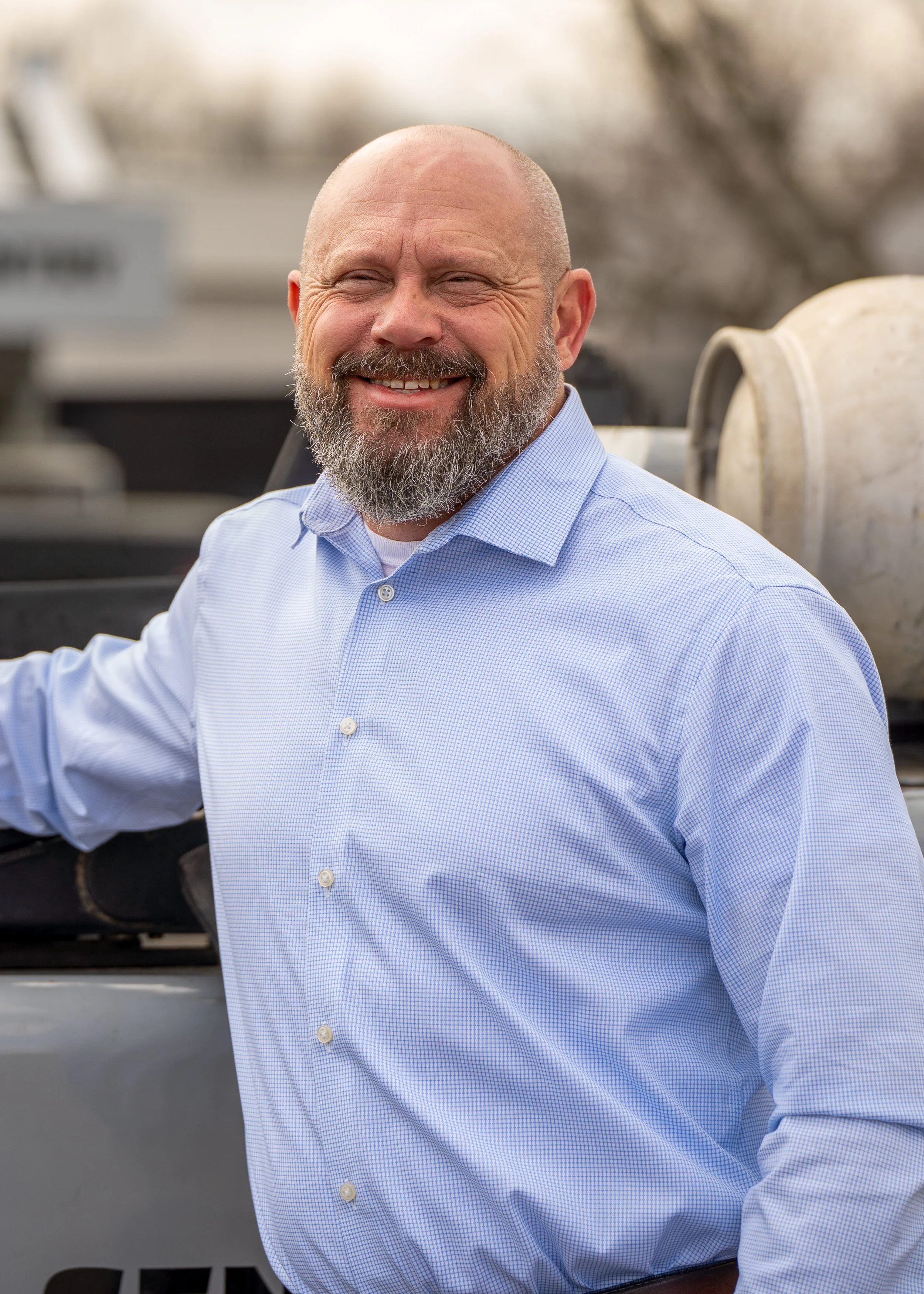 A smiling man with a beard in a light blue checkered shirt, standing outdoors near large barrels or drums.