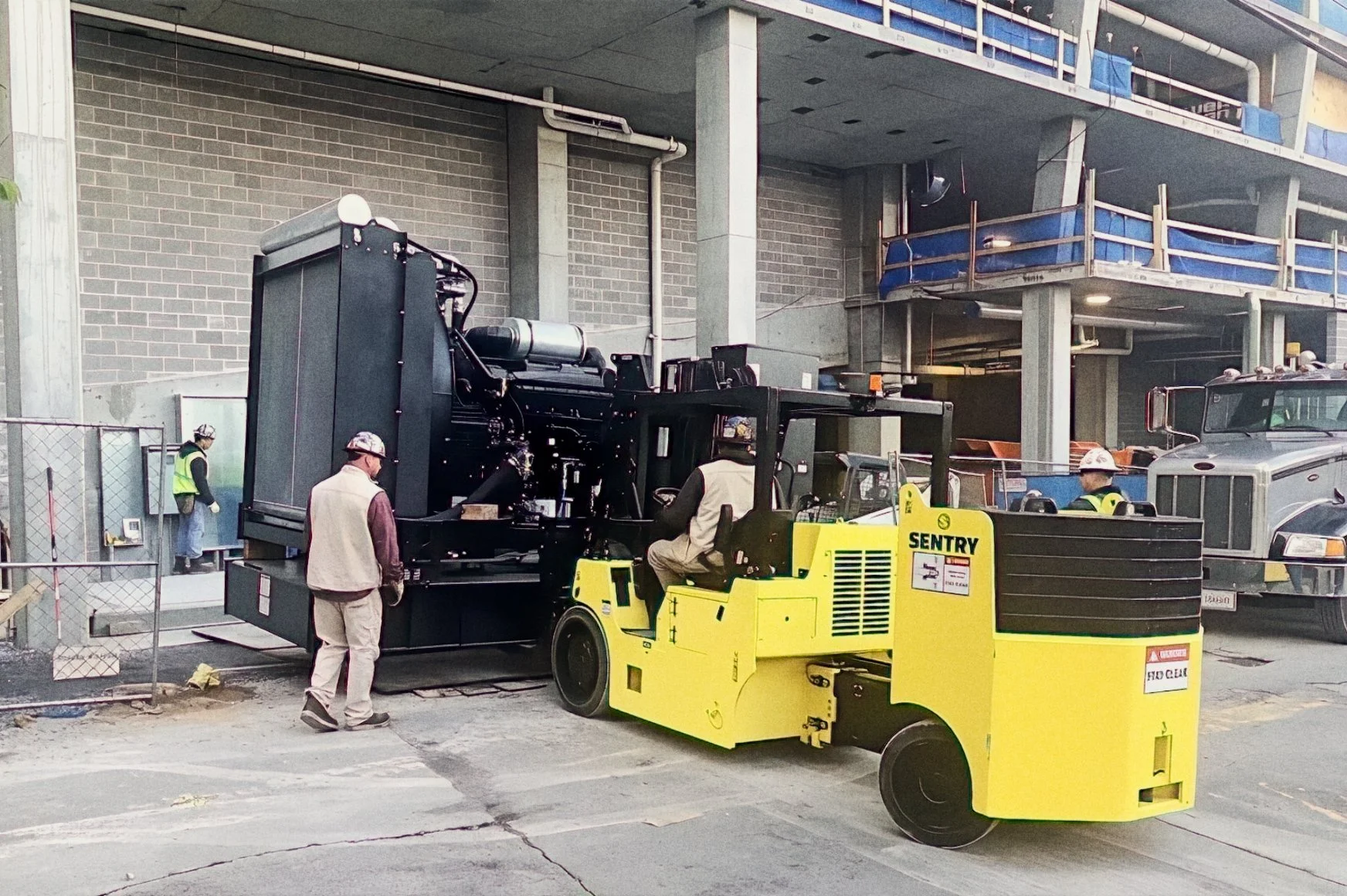 Construction workers and equipment on a building site, with a large black machine, a yellow industrial mower, and a grey truck in the background, and a multi-story parking garage structure.