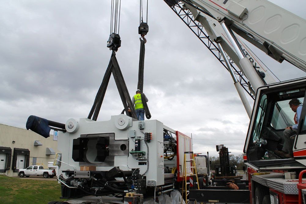 Construction workers operate a large mobile crane lifting a massive piece of equipment, with a building and parked vehicles in the background.