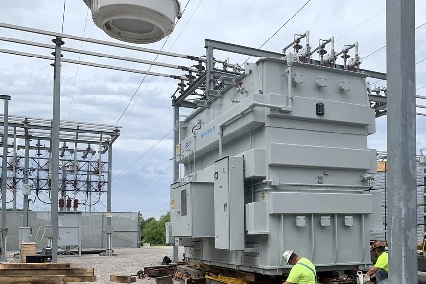 Large electrical transformer at an electrical substation, with workers in safety vests and helmets nearby.