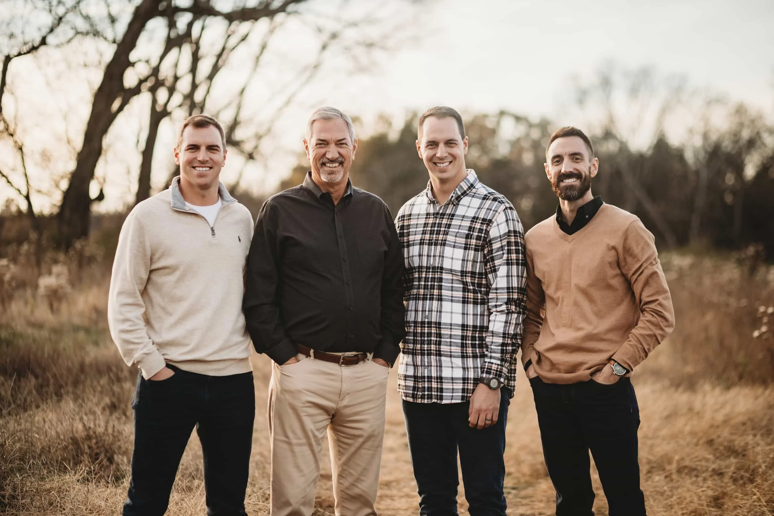 Group of four men standing outdoors in a field with trees in the background, smiling at the camera during sunset.