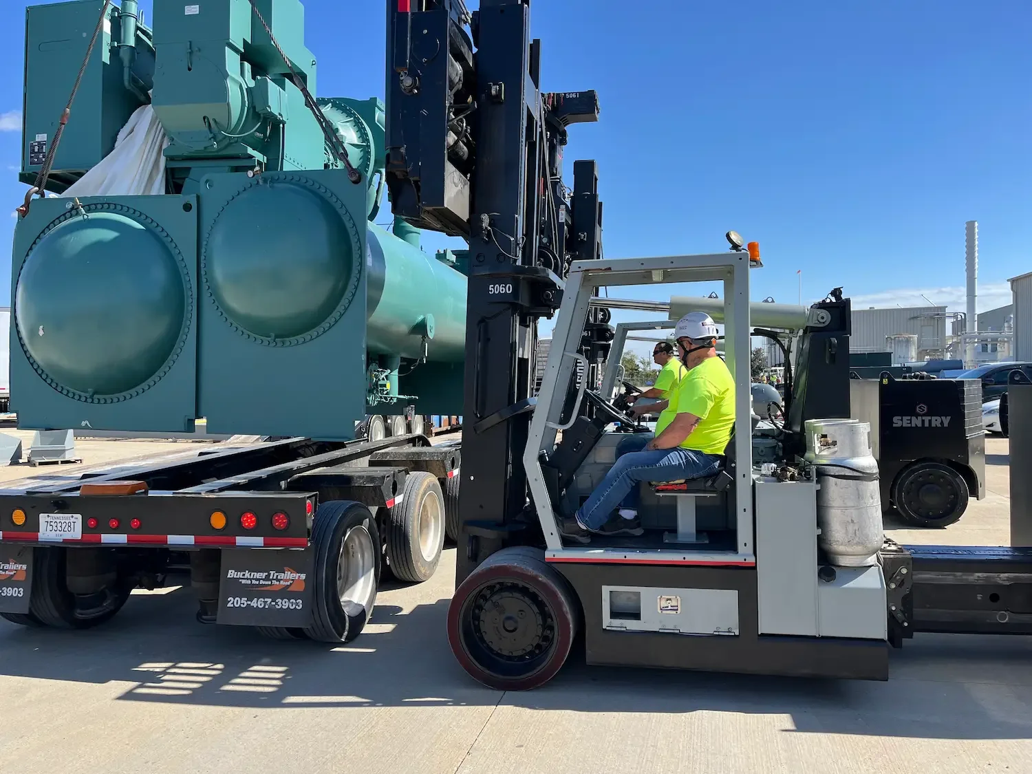 Workers using heavy forklift to unload large teal industrial unit from trailer