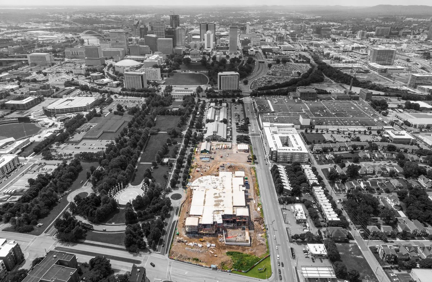 Aerial view of a construction site with buildings being worked on, surrounded by trees, roads, and city skyscrapers in the background.
