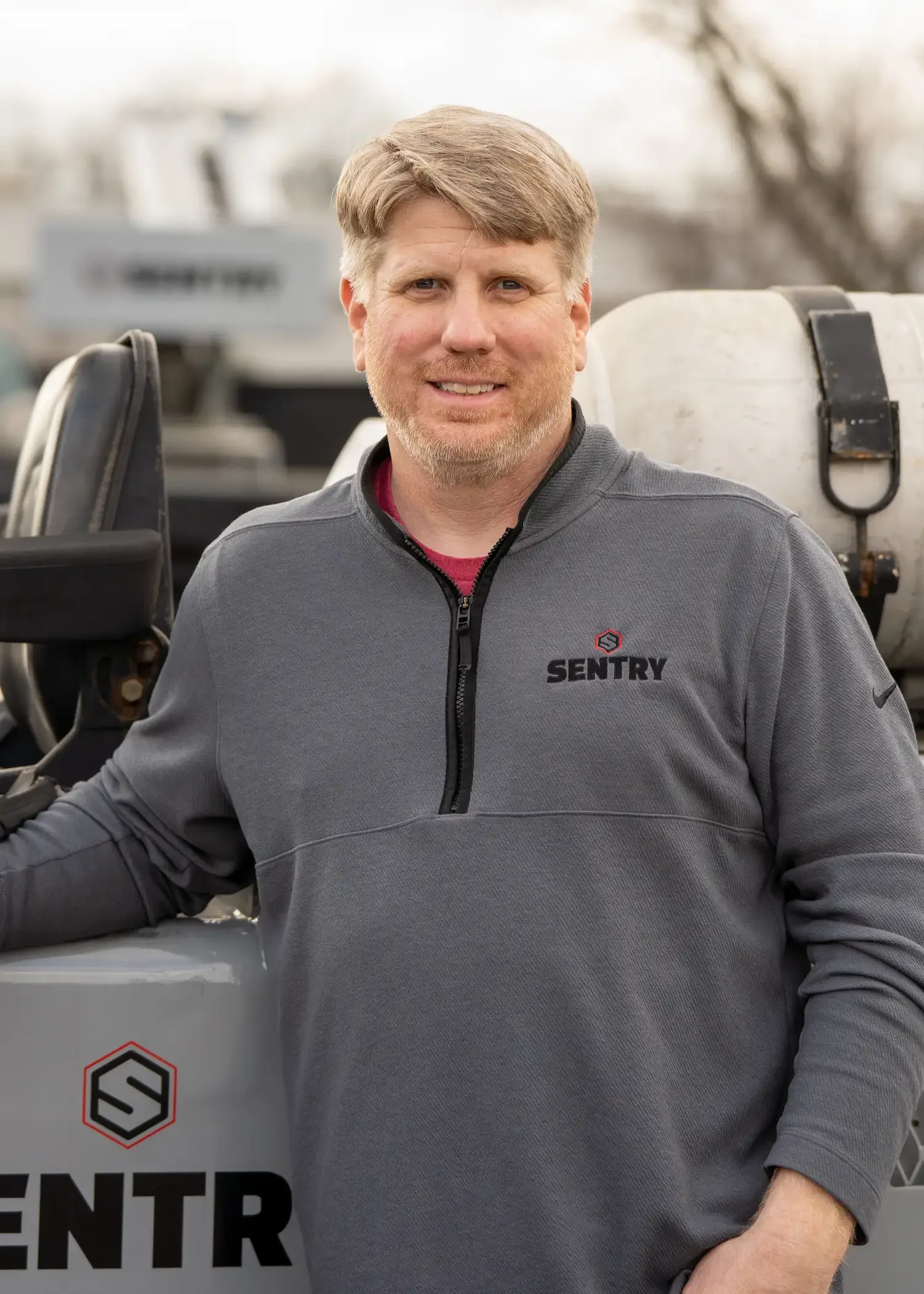 A man with light brown hair and a beard, dressed in a gray zip-up jacket with a logo and text, standing outdoors near a vehicle with a logo and partial word visible.