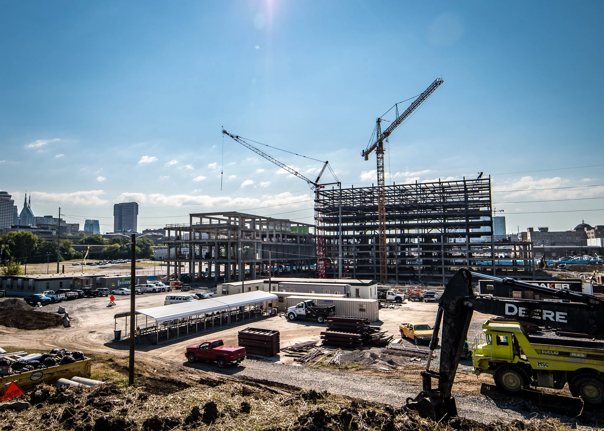 Construction site with cranes, partially built structures, and parked vehicles under a sunny sky.