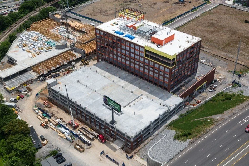 Aerial view of a multi-story building under construction next to a parking garage, with construction materials and equipment around, and a highway nearby.